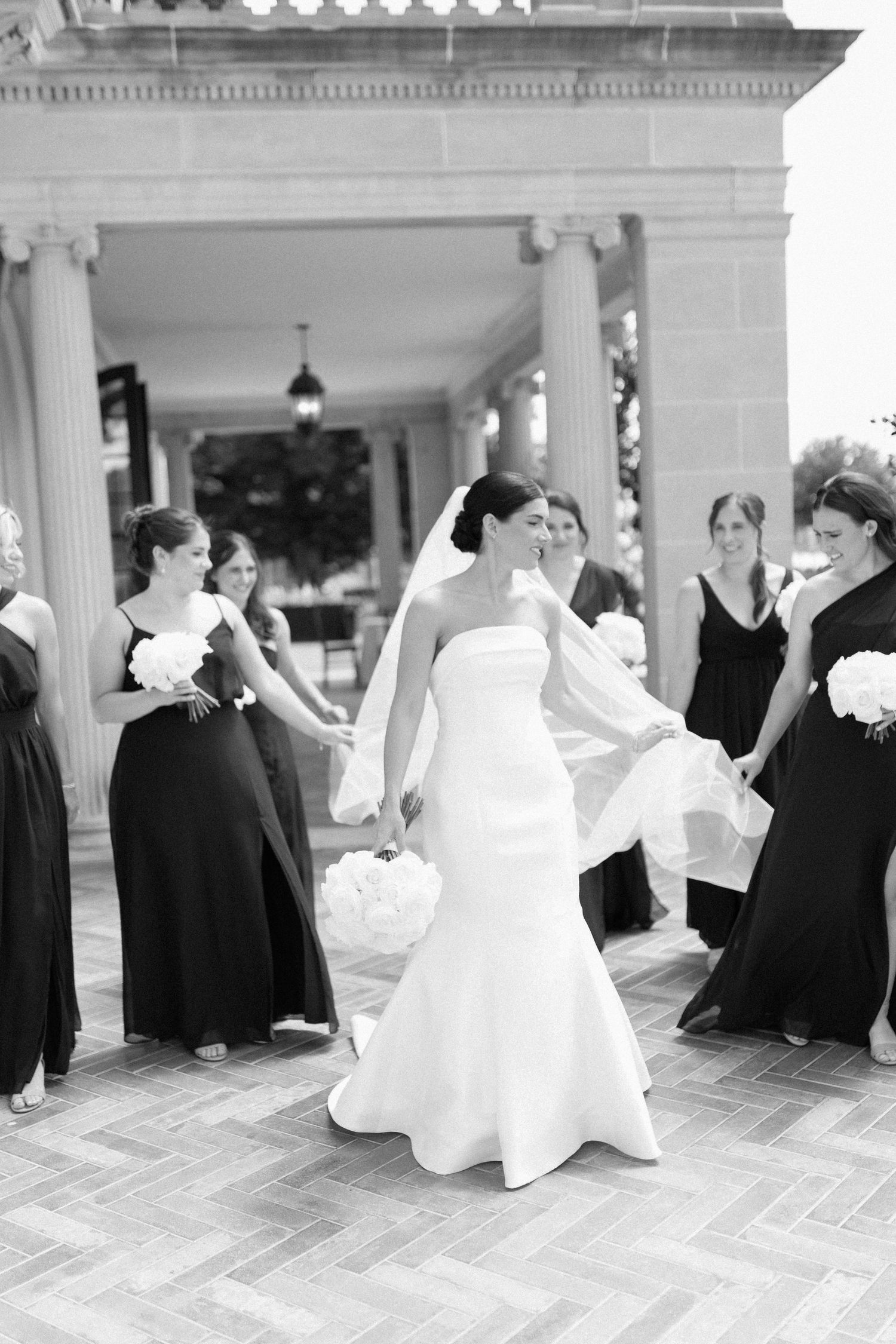 A wedding party in formal attire gathers on a columned porch with bridesmaids in black dresses.