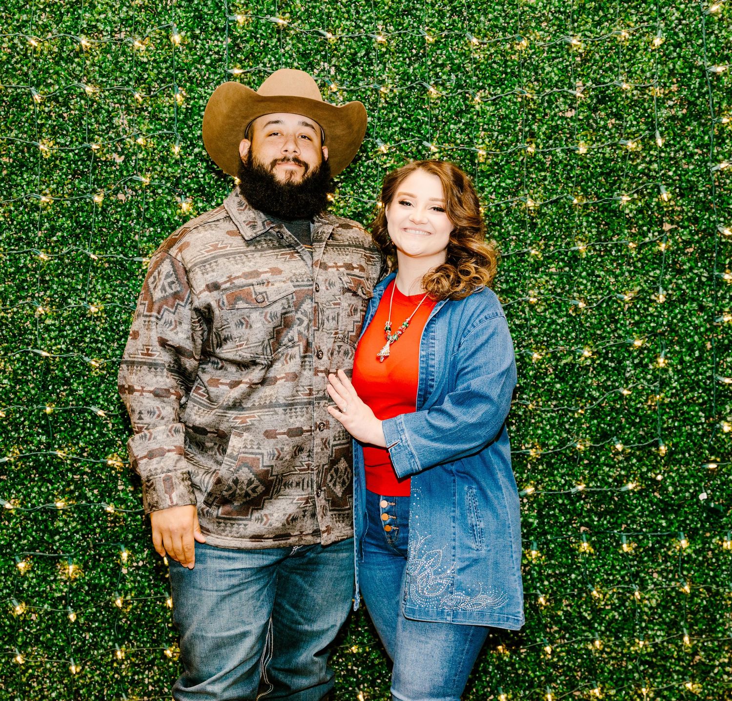 A couple poses together against a green floral backdrop, with one wearing camo and a cowboy hat and the other in denim.