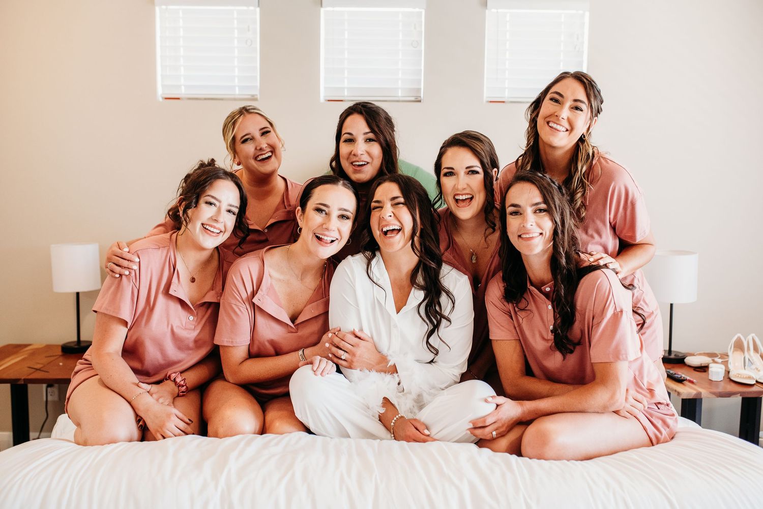 Group wearing matching pink robes sitting together on bed for bridal party photo.