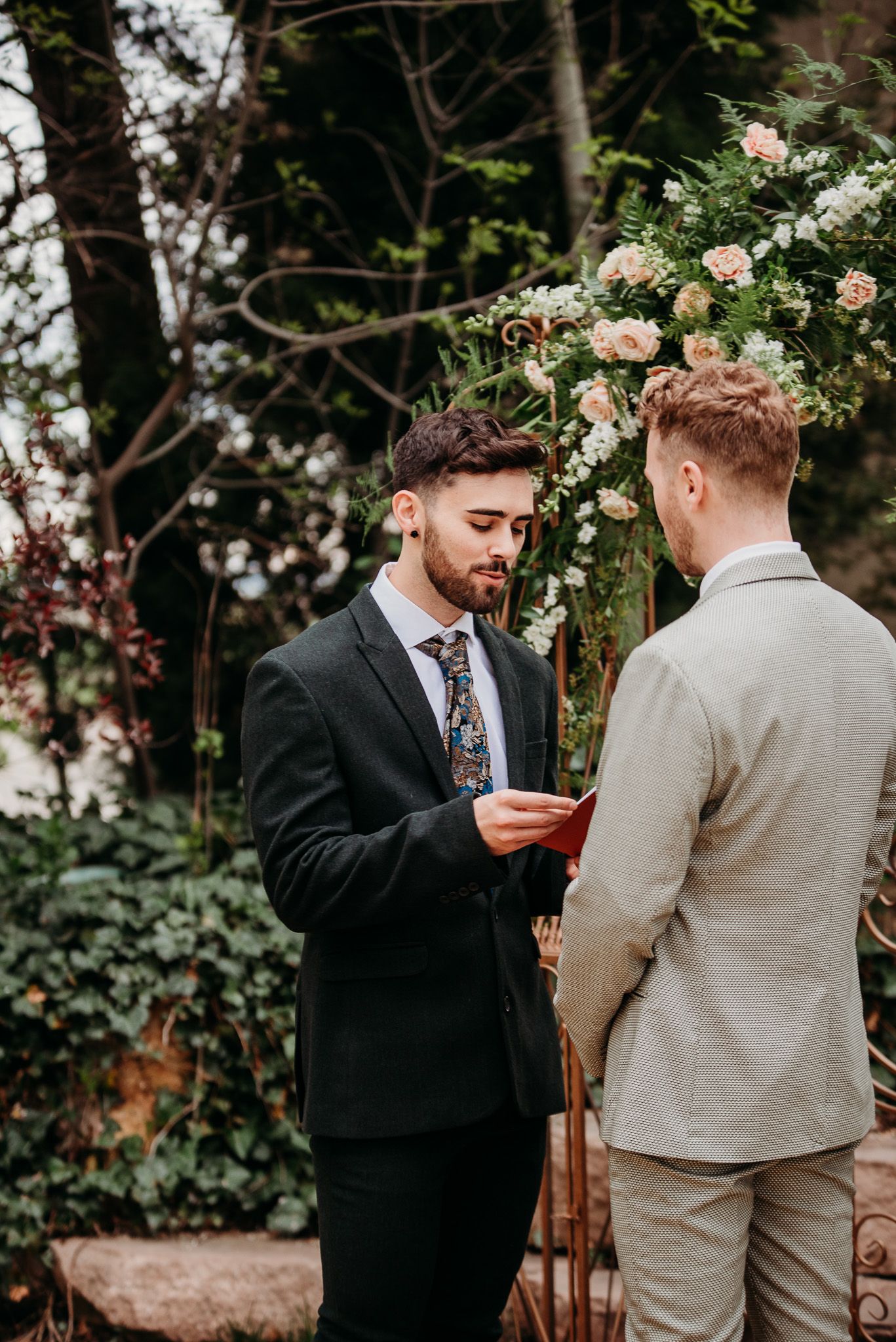 Two people in formal attire stand beneath a floral arch during an intimate outdoor ceremony.