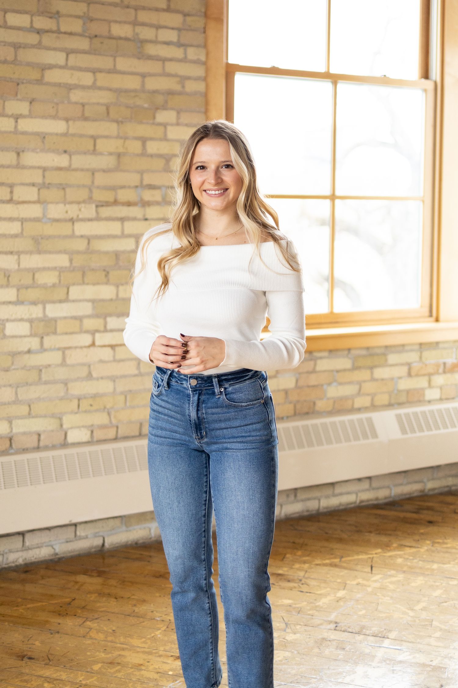 A woman in a white sweater and blue jeans stands smiling in front of a brick wall and window.