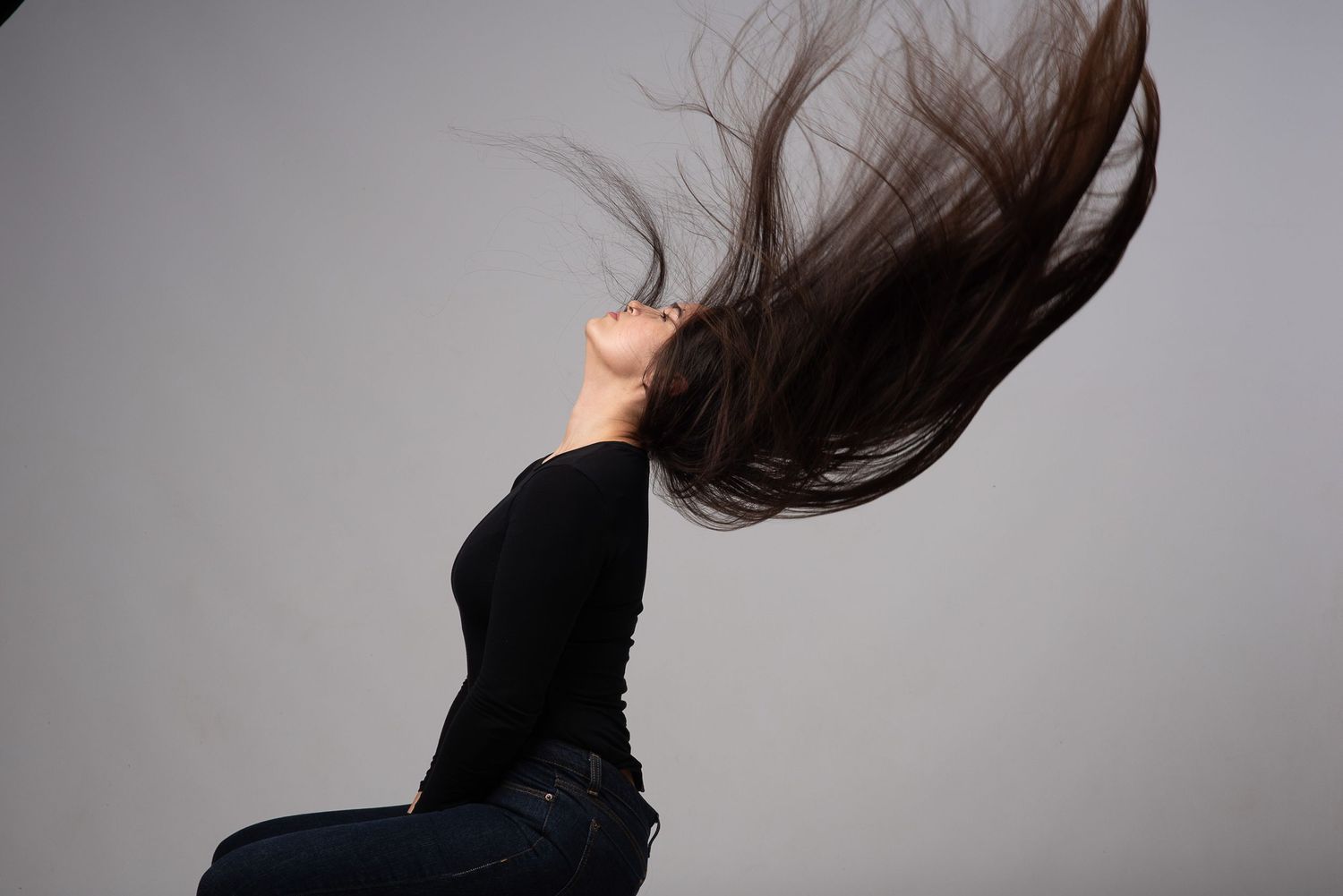 Long dark hair flips dramatically against a gray background as someone wearing black leans back in motion.