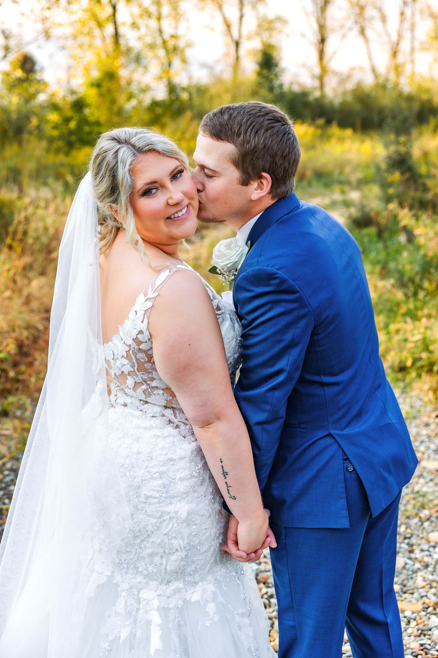 A romantic wedding photo showing a couple in a blue suit and lace wedding dress embracing in a golden sunlit field.