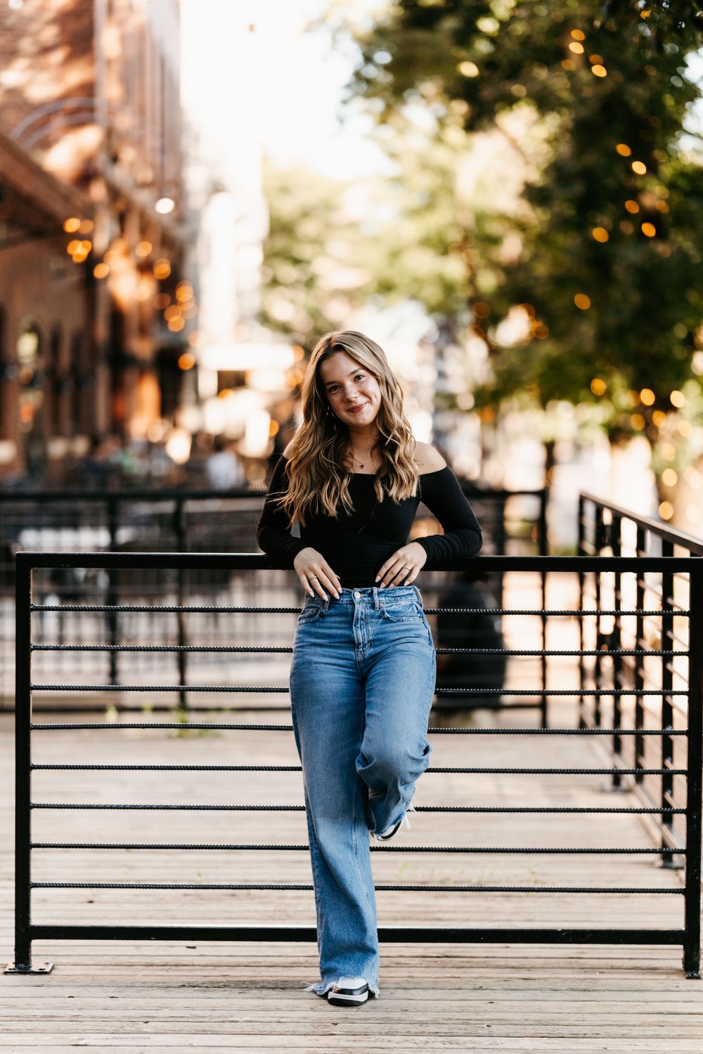 A casual outdoor portrait of someone in a black sweater and wide-leg jeans standing on a wooden walkway at sunset.
