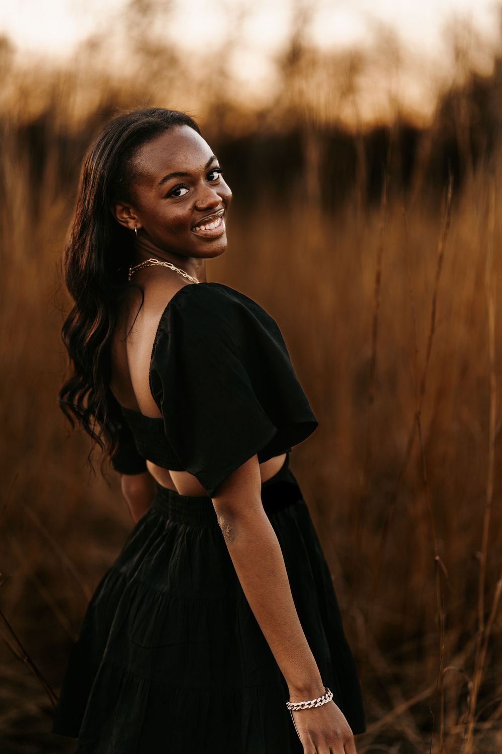 A joyful portrait in a black dress against tall grass at sunset.