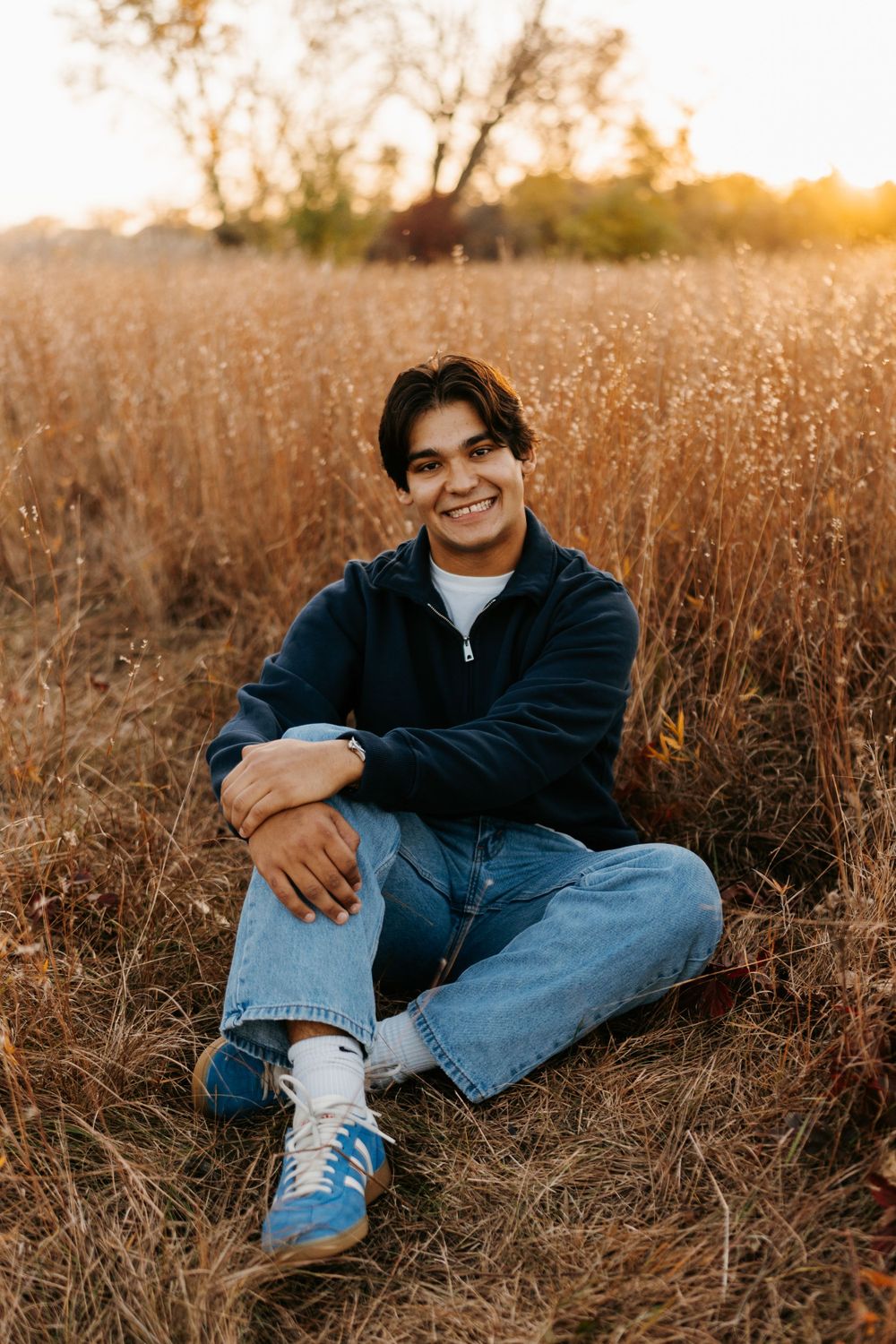 A casual portrait in denim and navy sweater sitting in golden grass at sunset.