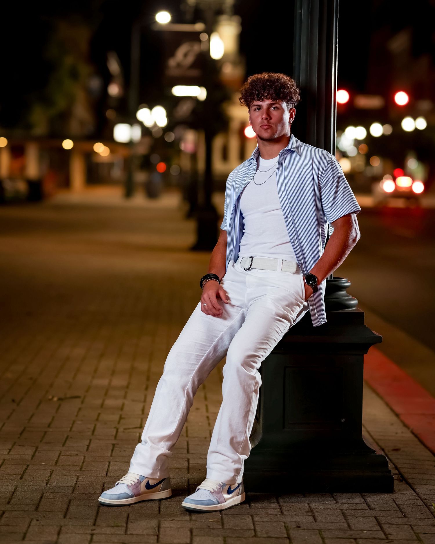 A person in all white casual attire leans against a post on a city street at night with bokeh lights in the background.