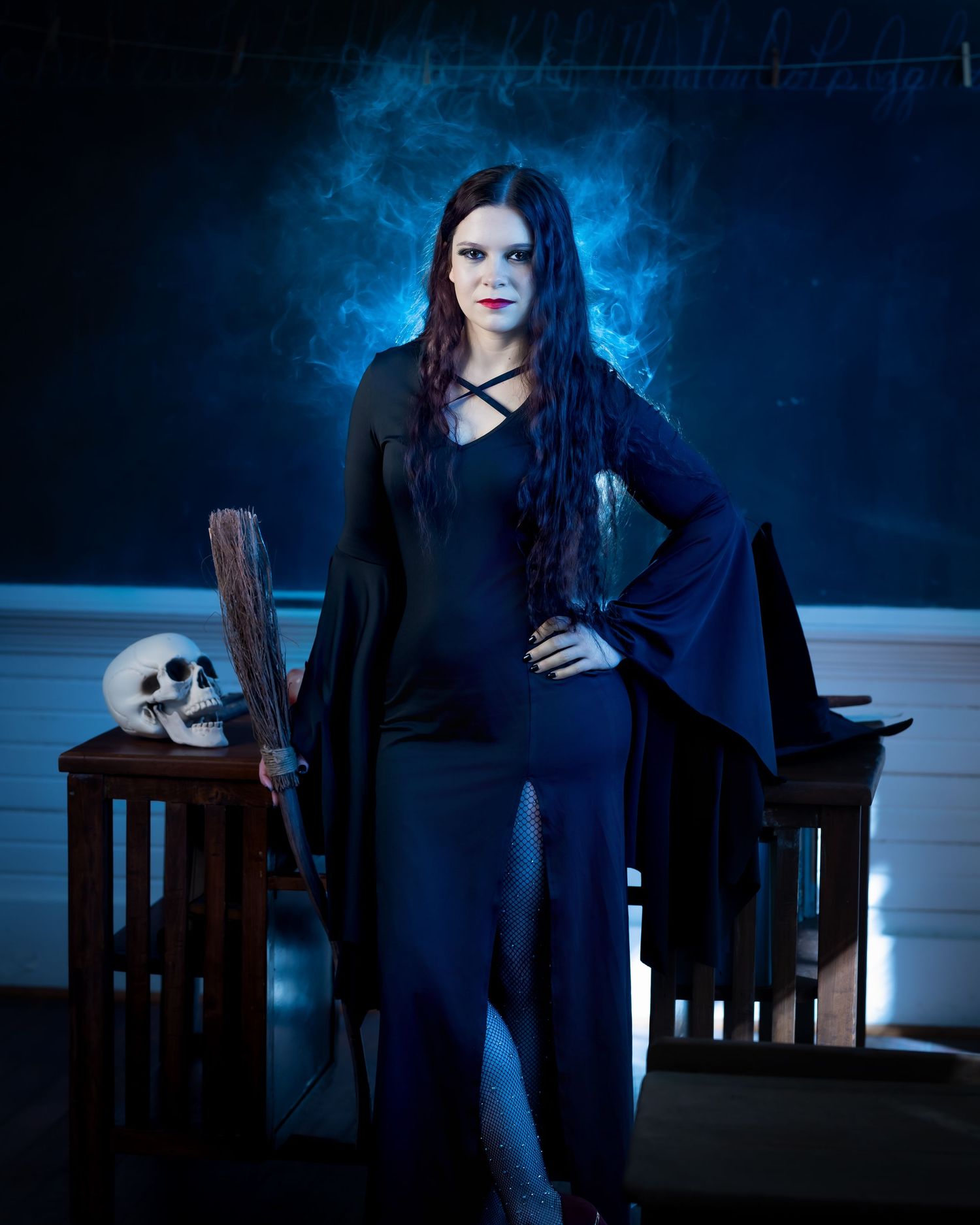 Gothic figure in black bell-sleeve dress poses with skull and lantern props against ethereal blue smoky background.