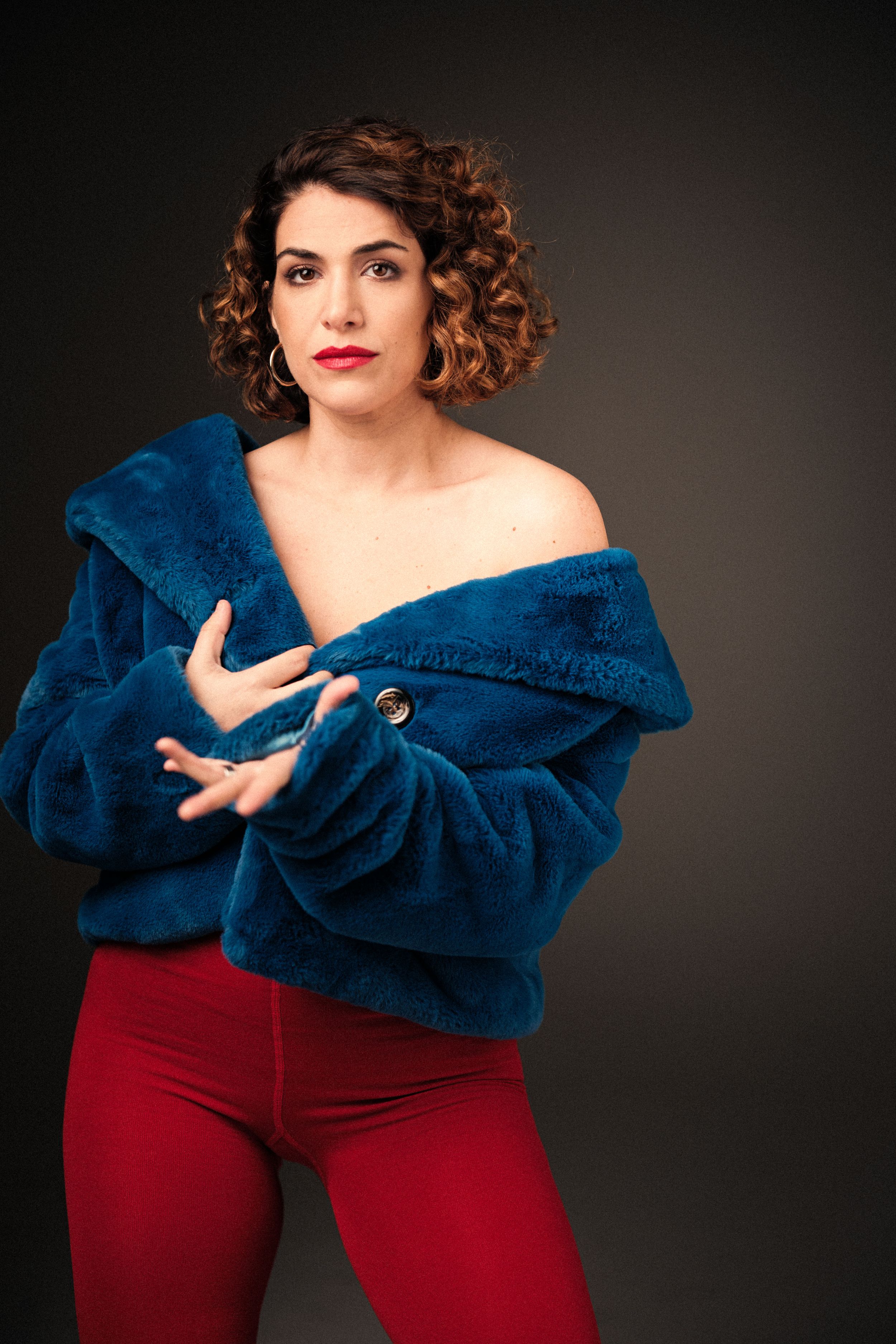 Expressive gestures while wearing plush blue jacket and red pants in studio setting.