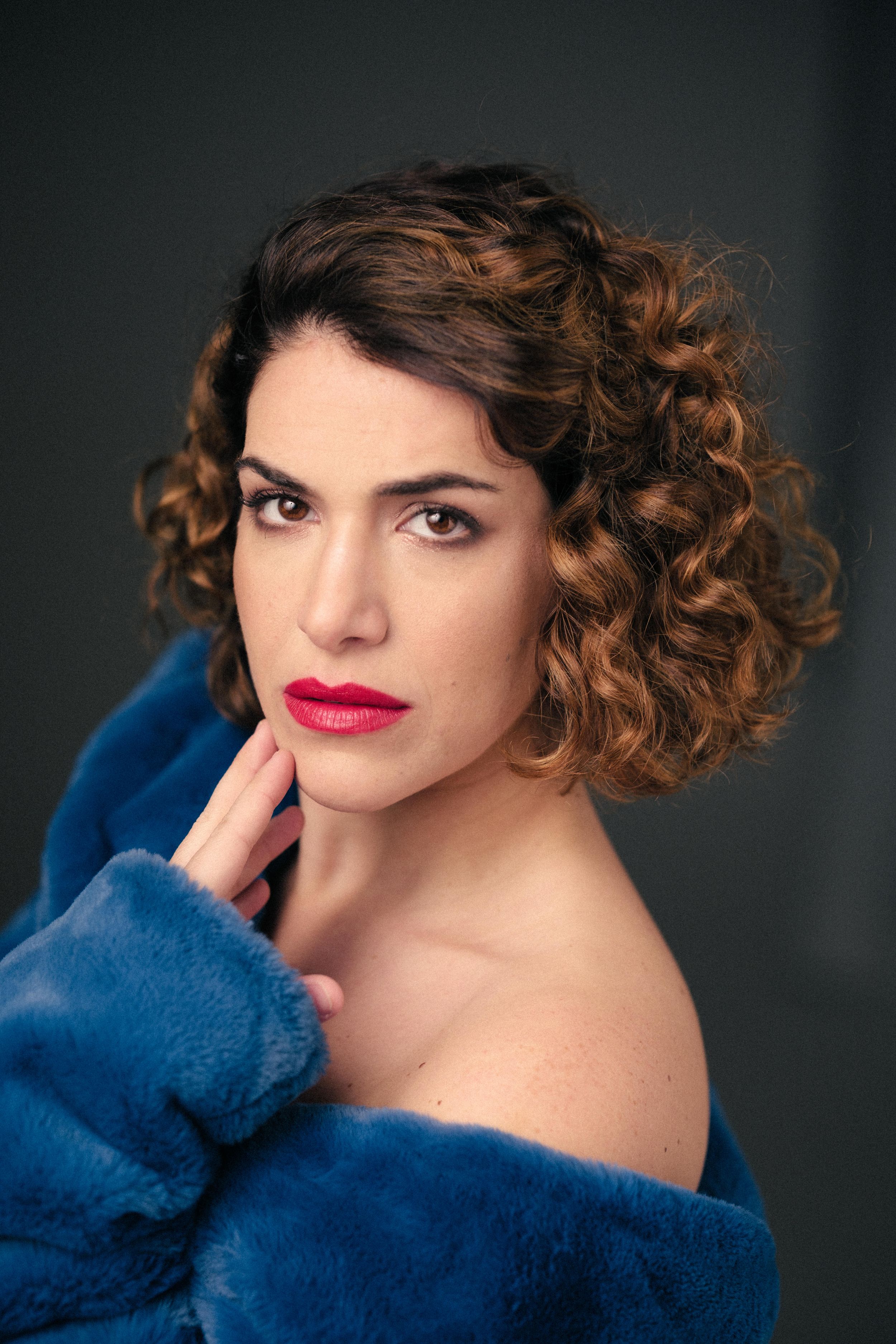 Glamour portrait with curly hair and blue fur coat against dark backdrop.