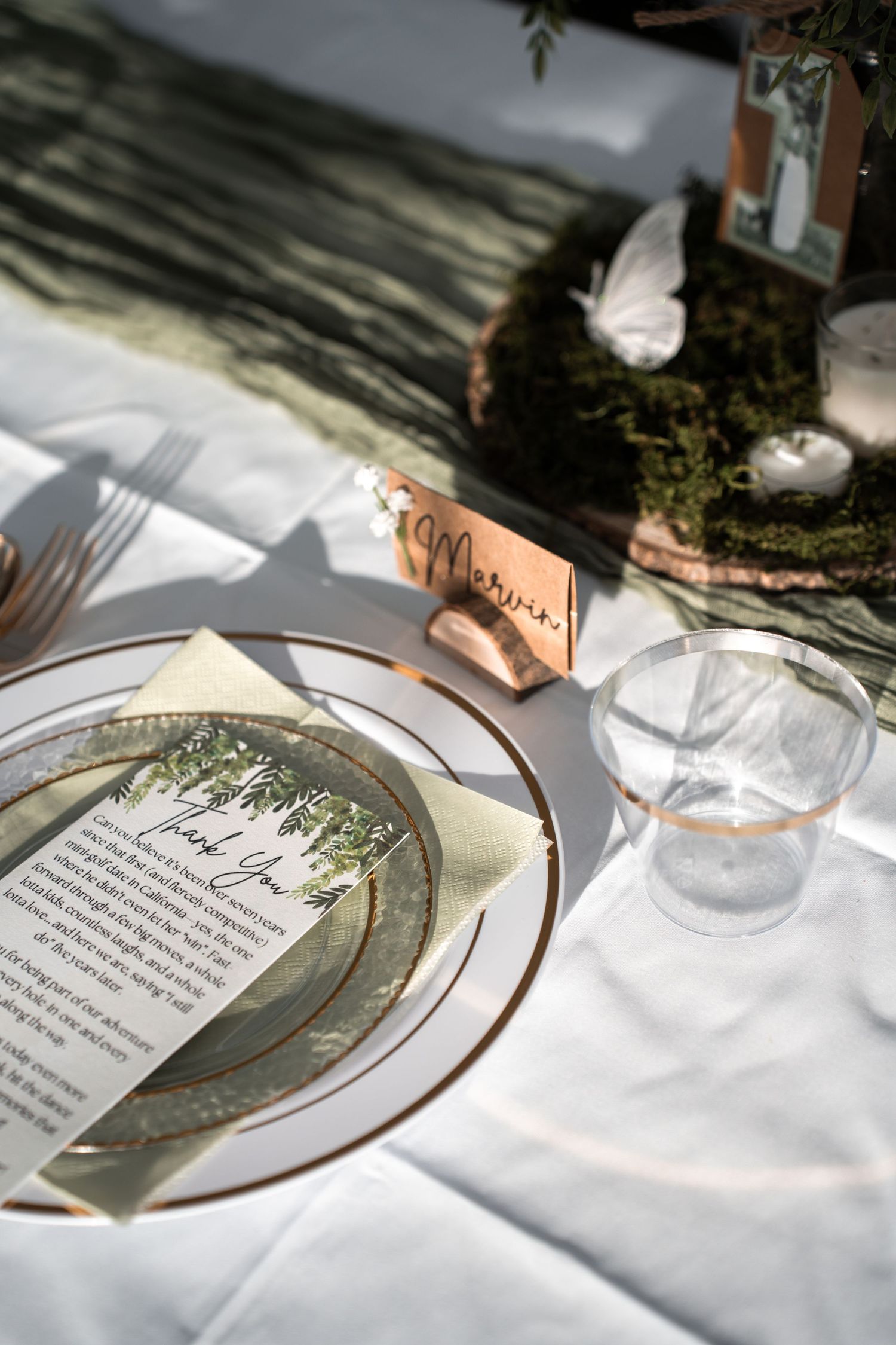 Place settings at a formal wedding reception with menus on decorative plates and sunlit white tablecloths.