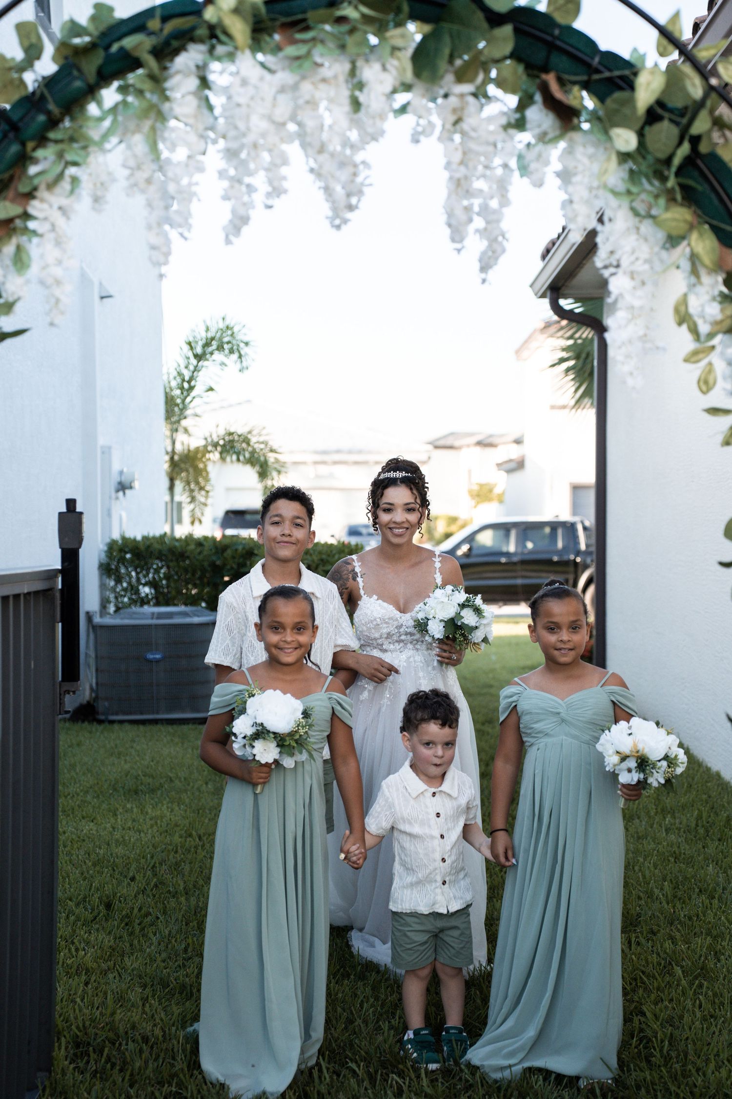 Bridal party in sage green dresses stands in a row under an elegant white floral arch outdoors.