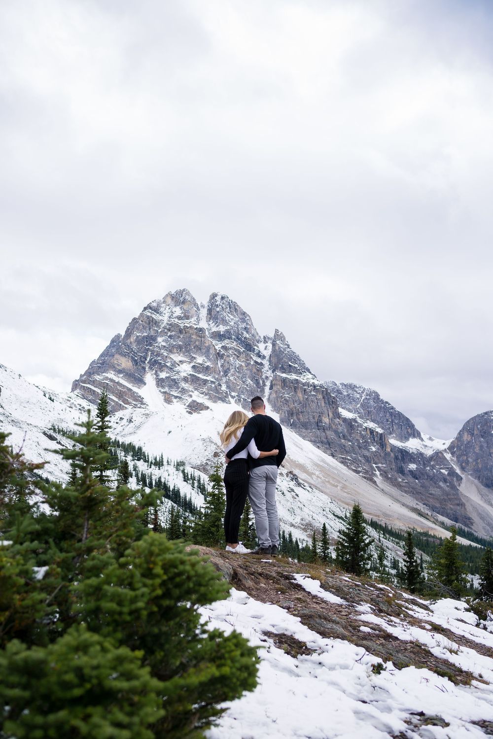 Snow-capped mountain peaks rise majestically behind evergreen trees in a dramatic winter landscape.