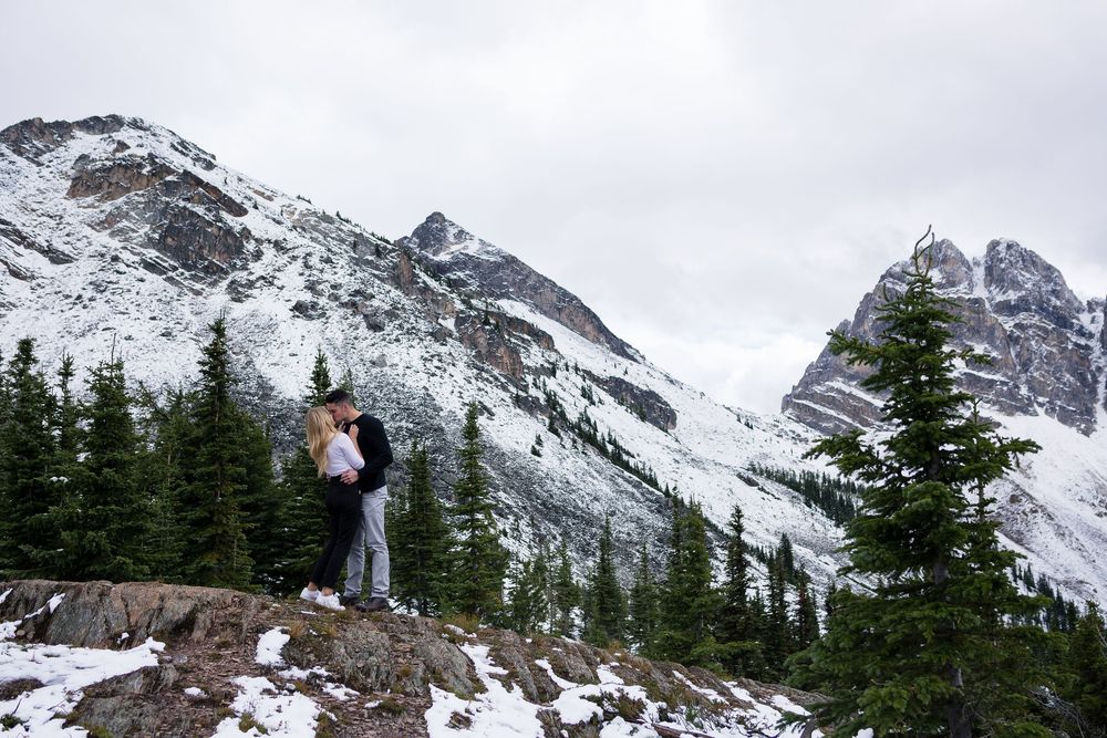 A couple poses at a scenic mountain overlook with snowy peaks and evergreen trees in winter.