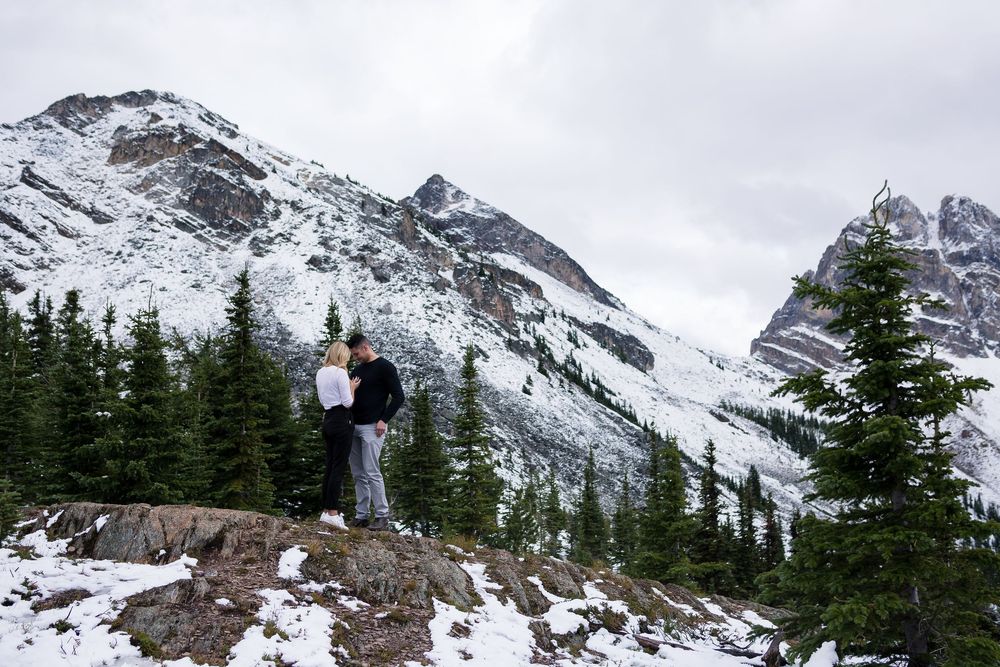Dramatic mountain landscape with snowy peaks and evergreen trees dotting the rugged rocky terrain under cloudy skies.