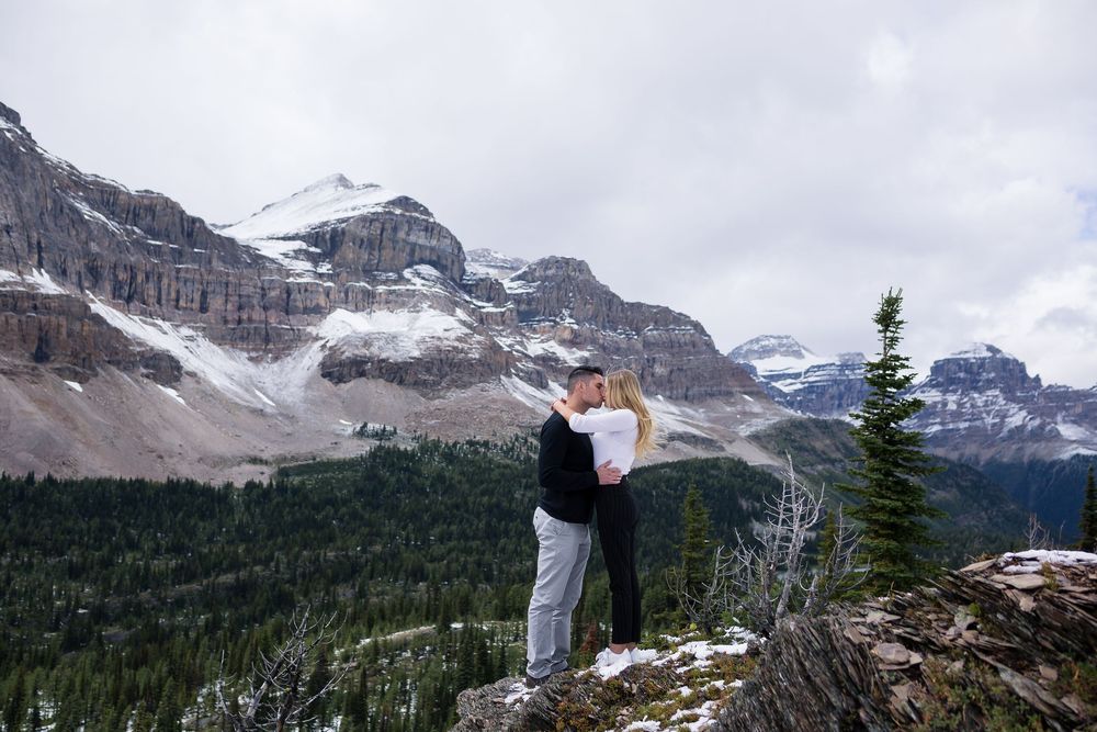 Person stands at mountain viewpoint looking over snow-capped peaks and dense forest in Canadian Rockies.