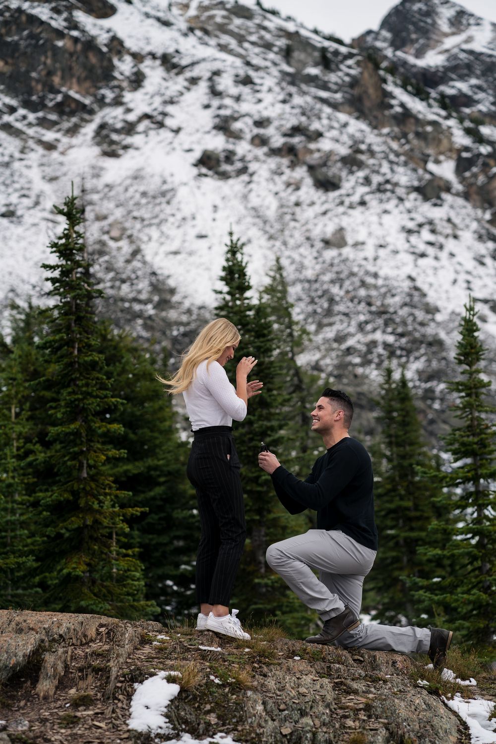 A couple share a romantic embrace on a snowy mountain ledge with evergreen trees and snow-capped peaks in the background.