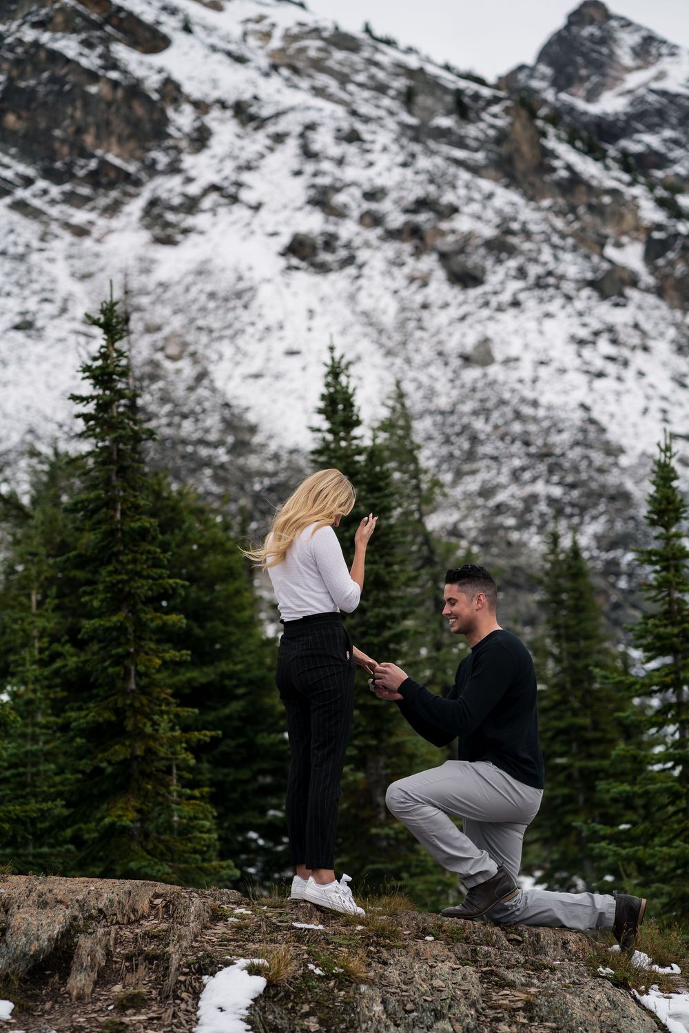 A couple shares a romantic moment on a snowy mountain ledge with evergreen trees and snow-capped peaks in the background.