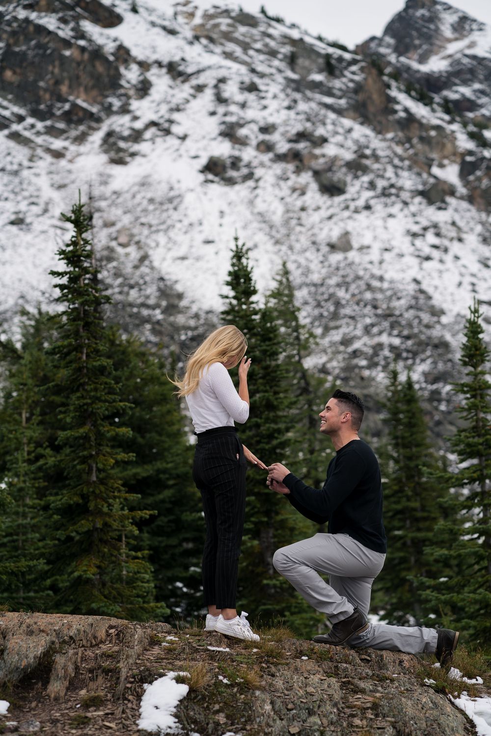A proposal unfolds on a rocky outcrop with snowy mountains and evergreen trees creating a stunning backdrop.
