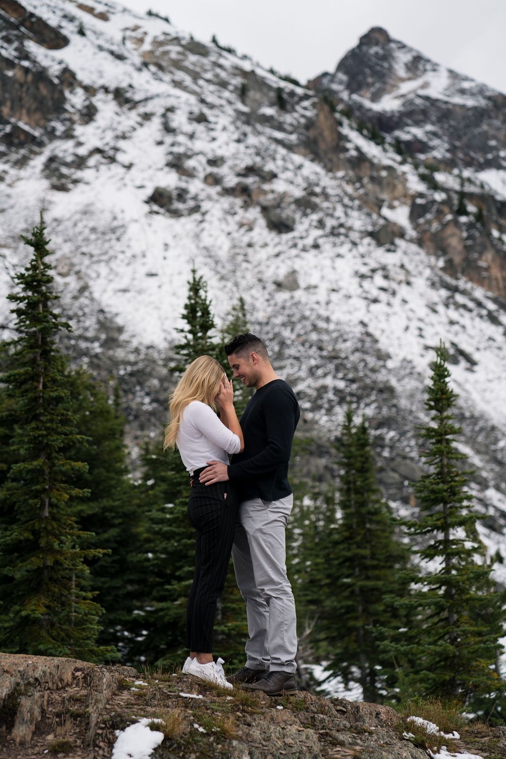 A joyful moment shared on a mountain ledge surrounded by snow-covered peaks and pine forest.