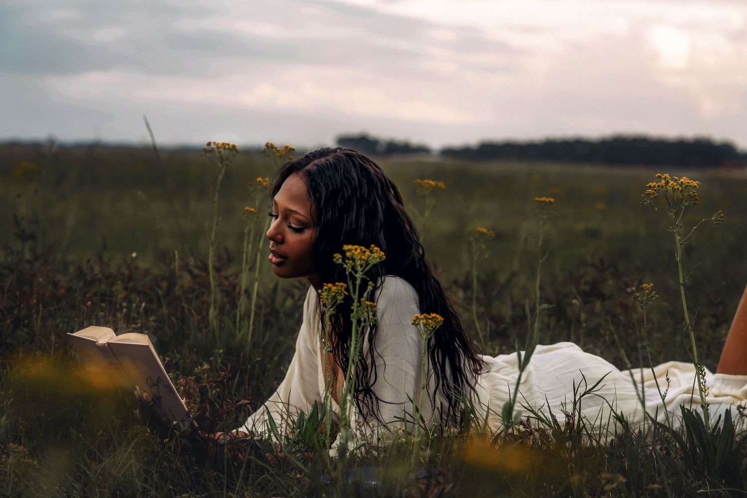 A Black woman with long braids lies in a wildflower field reading a book at dusk.