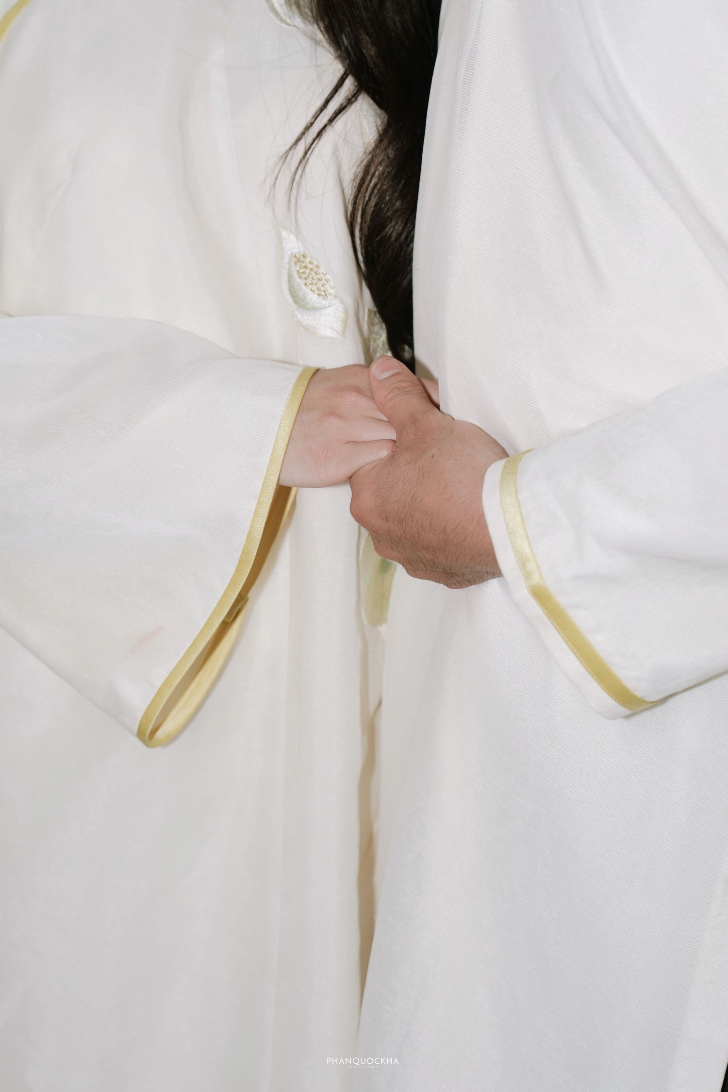 Close-up of Vietnamese couple in white and gold ao dai holding hands during traditional wedding ceremony.