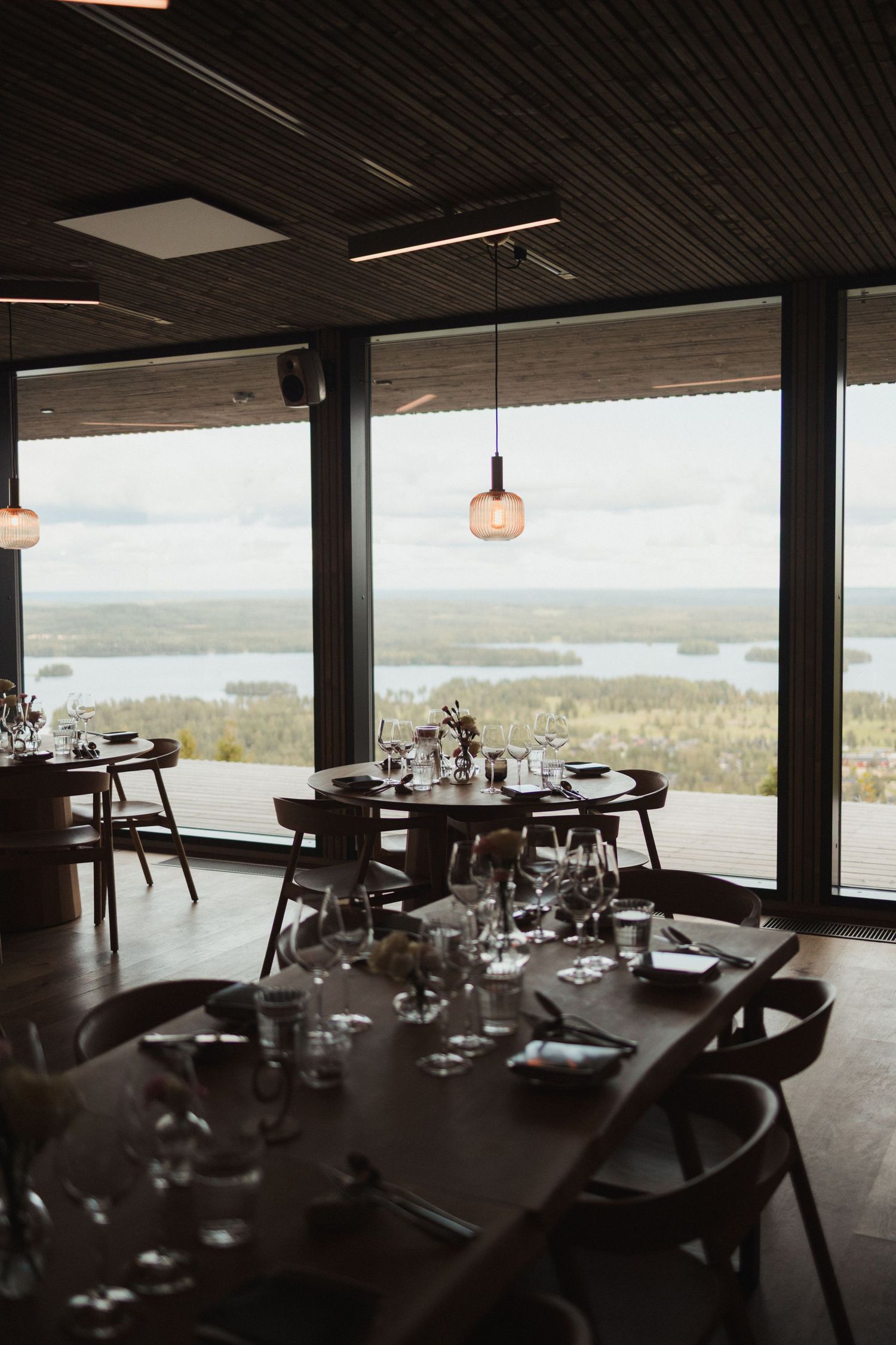 Modern restaurant dining room with panoramic windows overlooking a scenic lake and forest landscape.