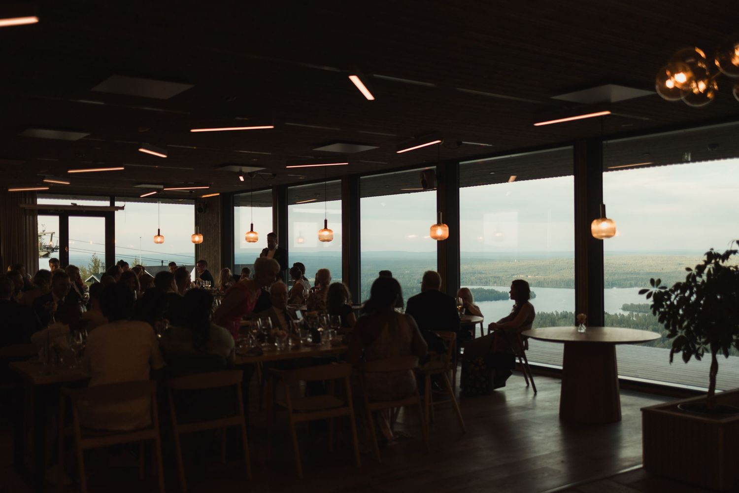 Restaurant interior at sunset with diners enjoying panoramic ocean views through floor-to-ceiling windows.