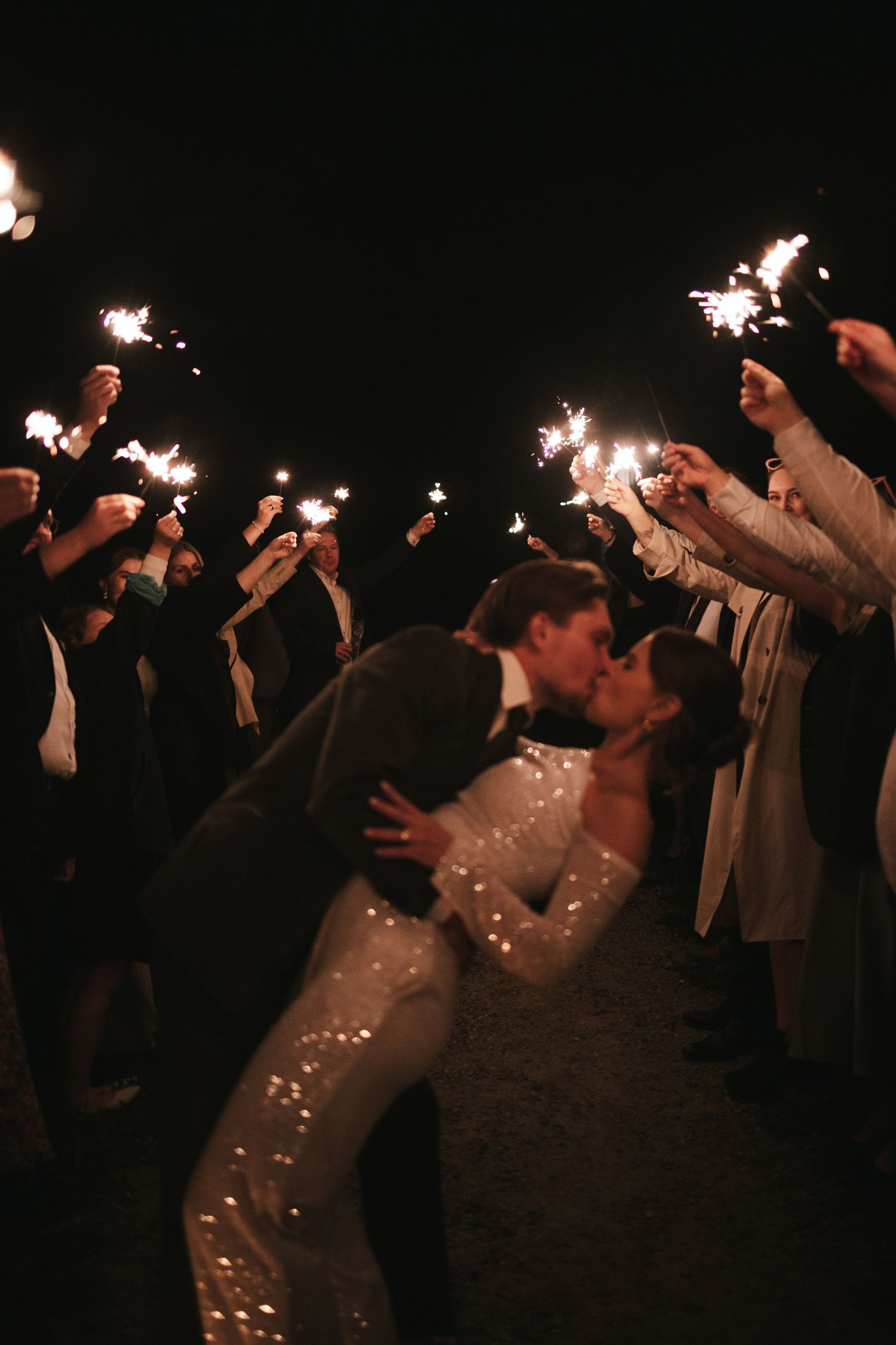 Newlyweds sharing a romantic sparkler moment surrounded by guests at night.