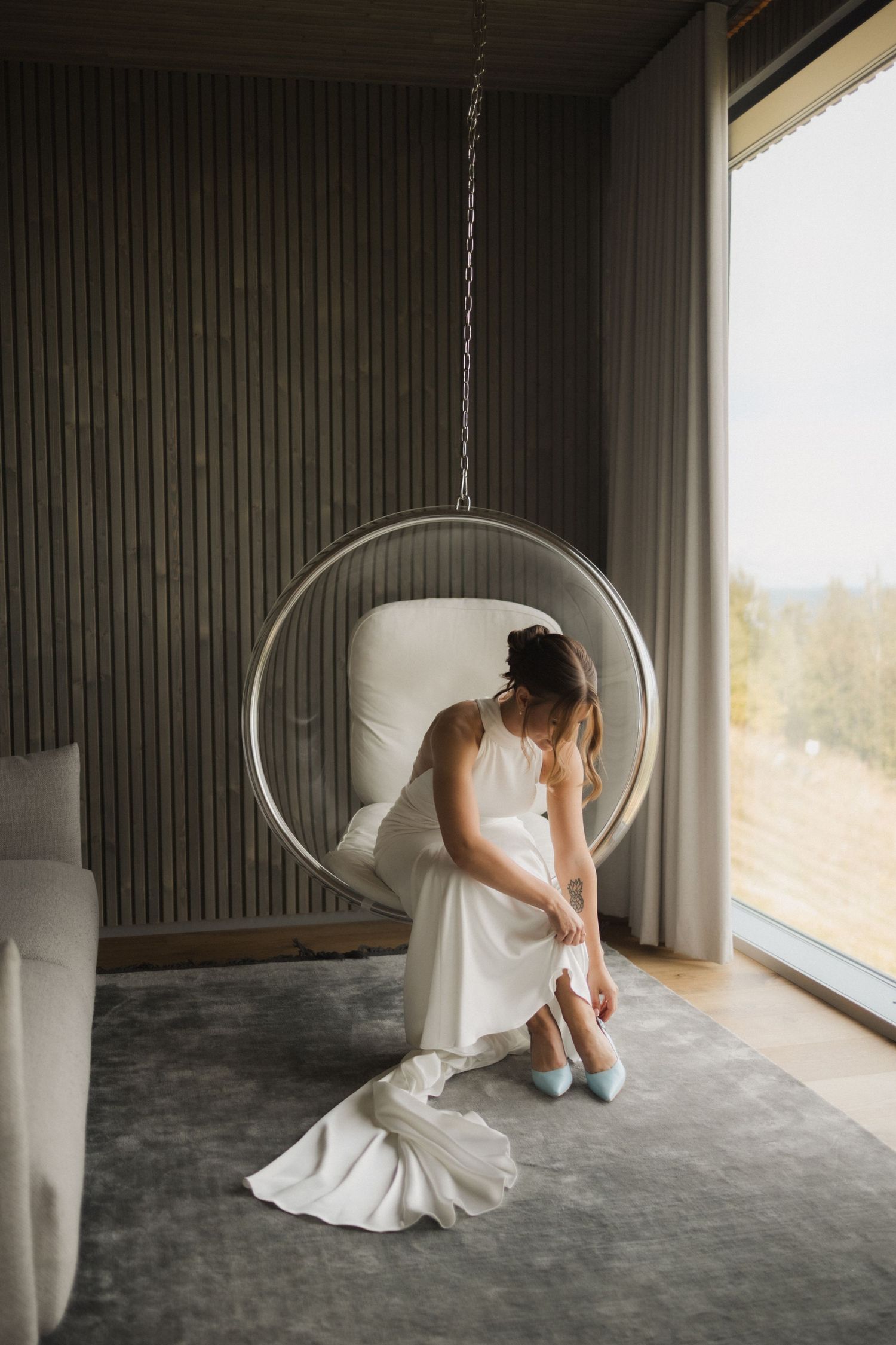 A white wedding dress flows elegantly as someone sits in a suspended bubble chair against a modern interior.