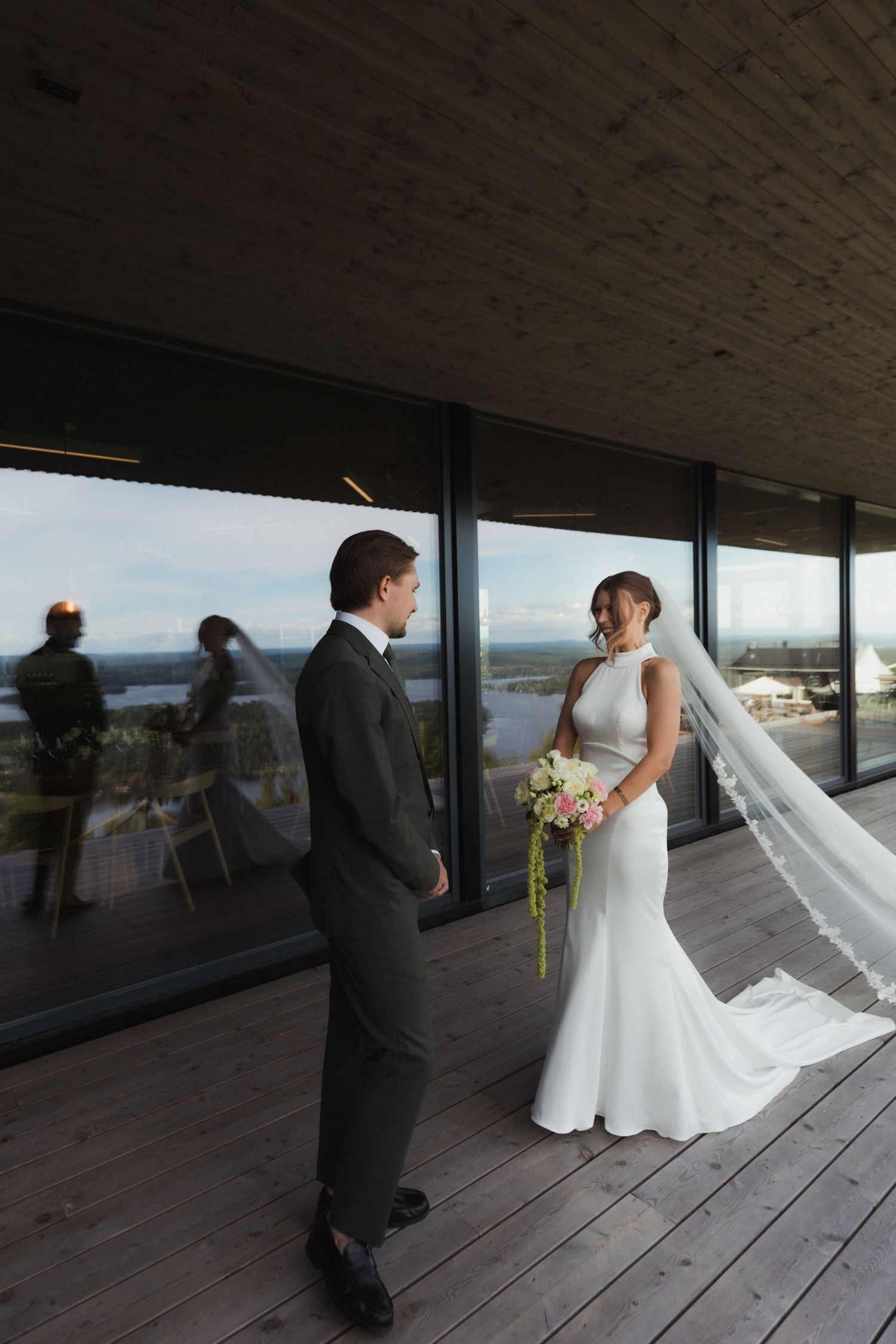 A wedding couple stands on a covered deck overlooking a scenic waterfront view.