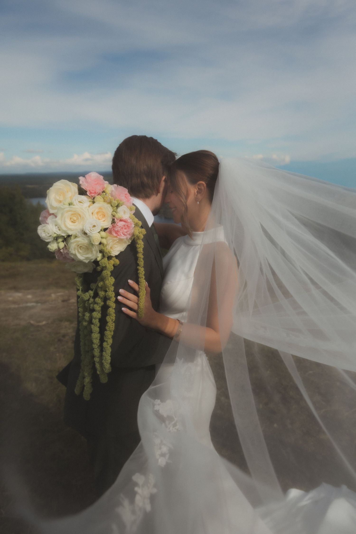 A wedding couple with flowing veil and bouquet against a scenic mountain backdrop.