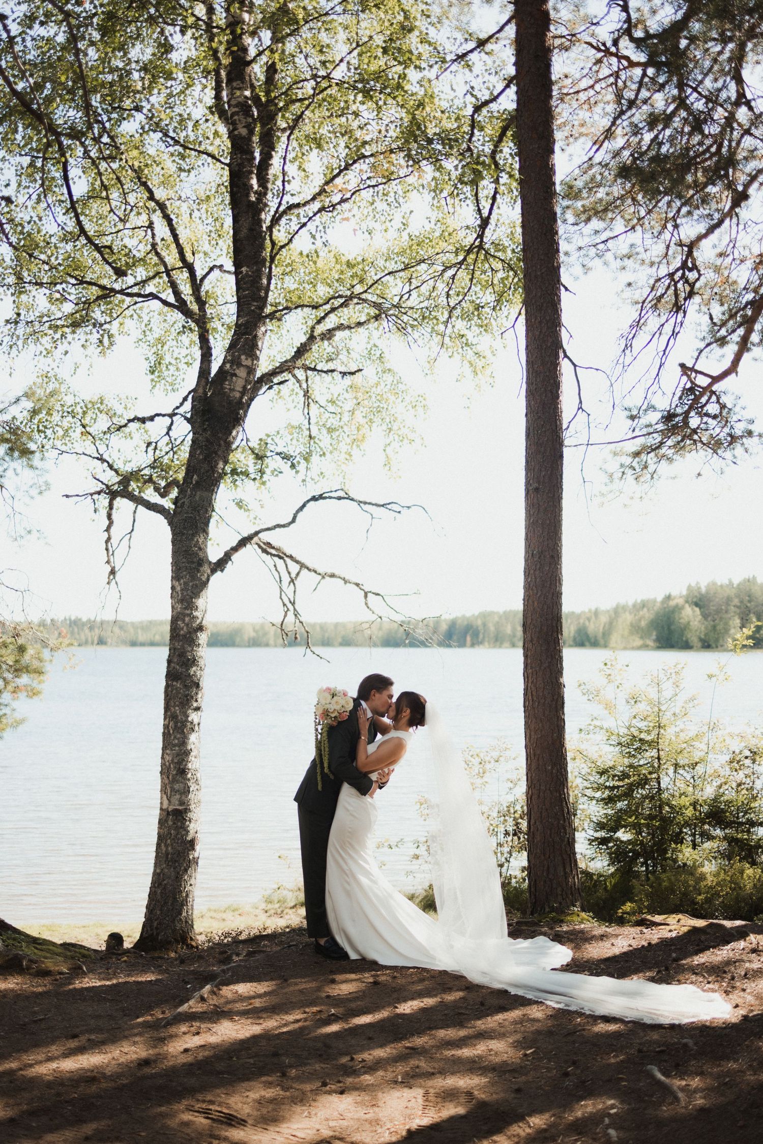 Wedding couple poses among tall pine trees by a serene lakeside on a sunny afternoon.