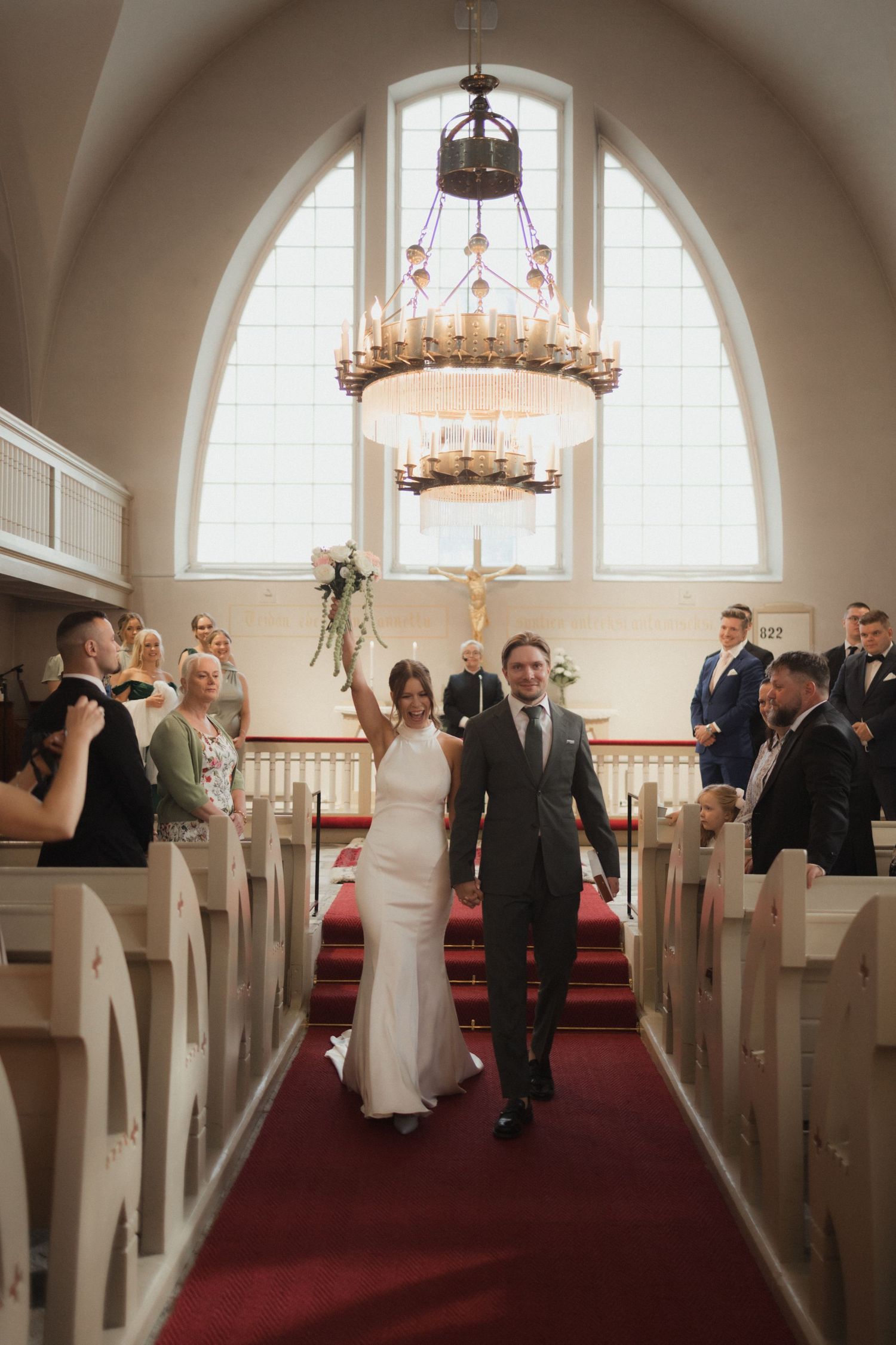 Wedding party exits down the church aisle after ceremony under ornate chandelier lighting.