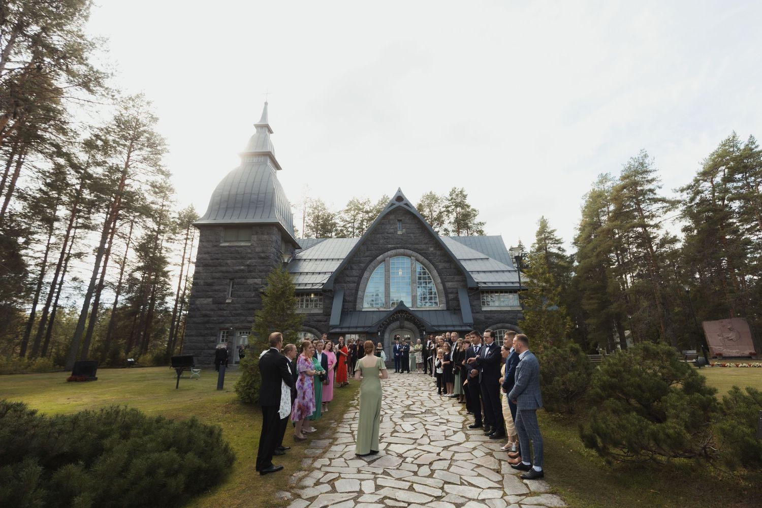Stone pathway leading to a chapel surrounded by pine trees and wedding guests.