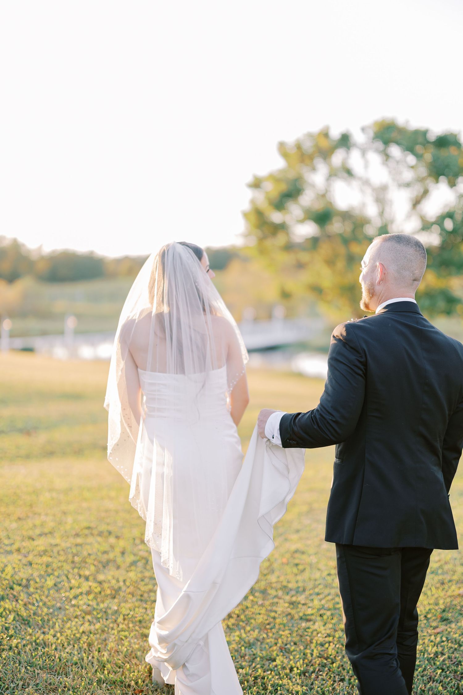 A romantic couple walking together at sunset in a grassy field with the bride wearing a flowing veil and white dress.