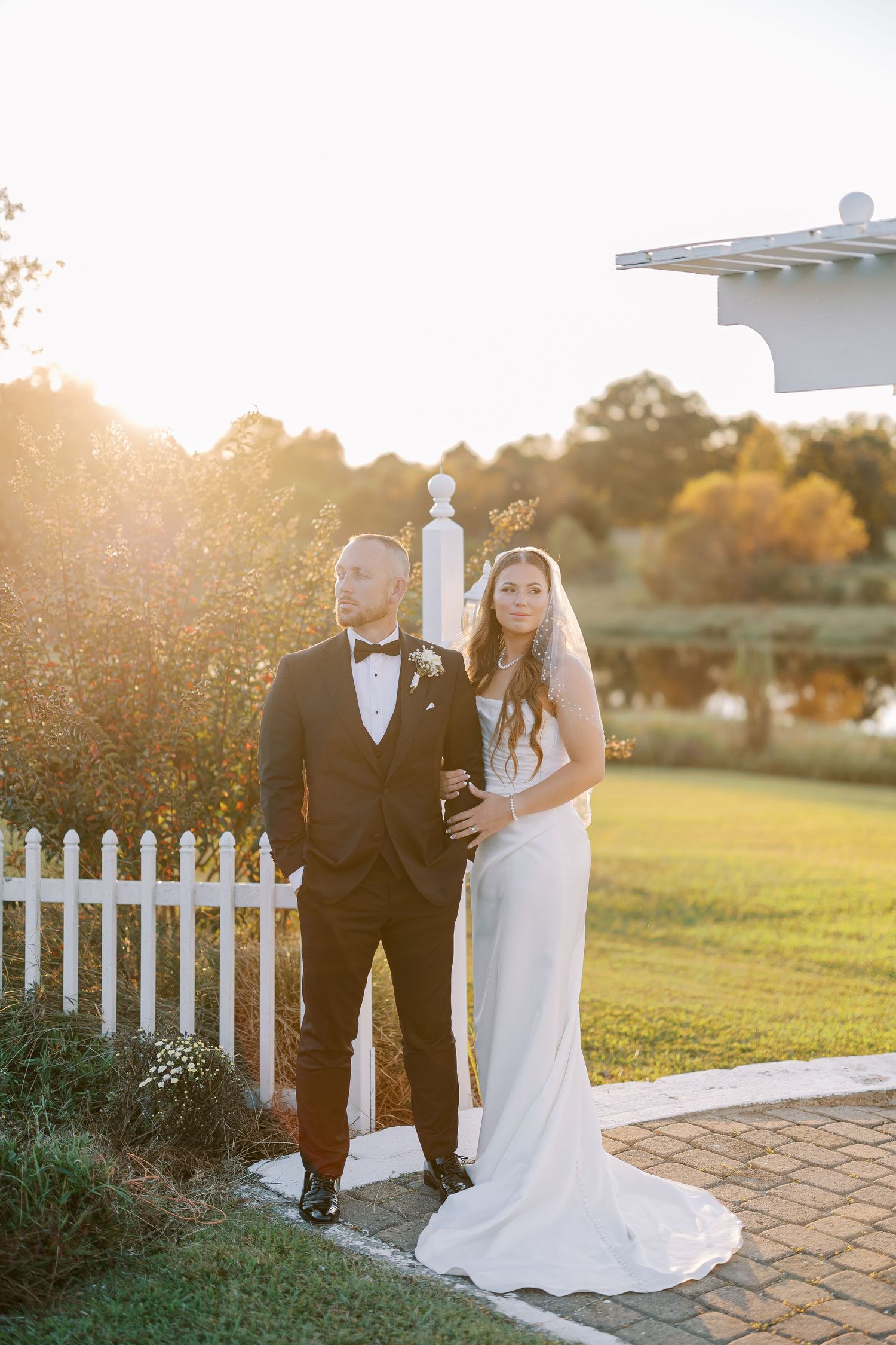 Couple poses together by white picket fence during golden hour sunset at their outdoor wedding ceremony.