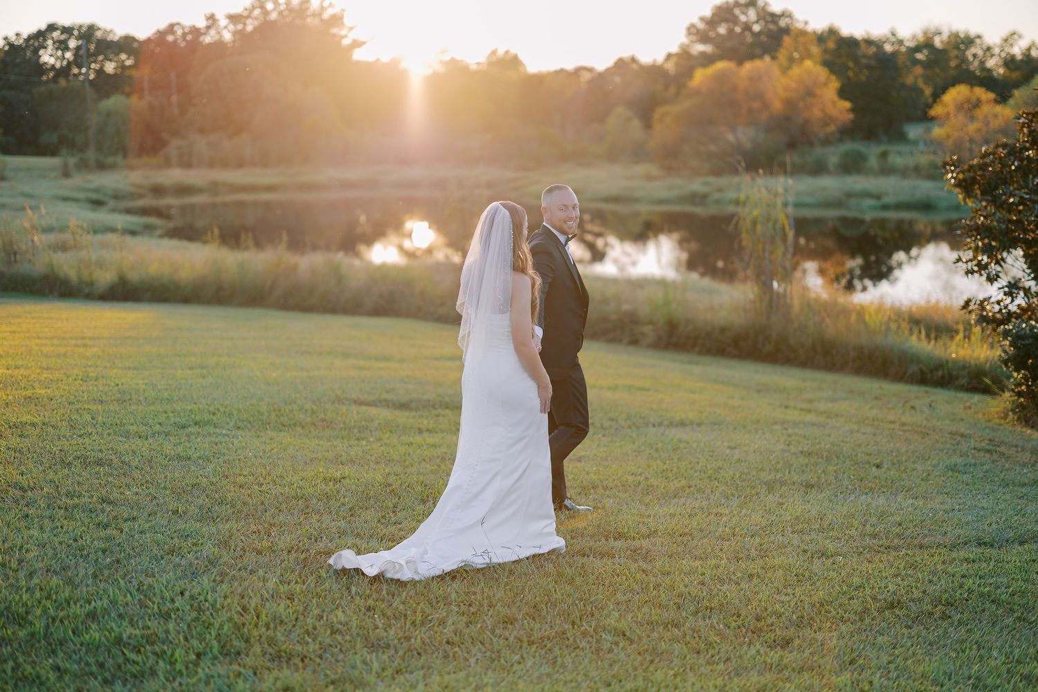 A romantic couple in formal wedding attire embraces during golden hour in a scenic outdoor setting near a lake.