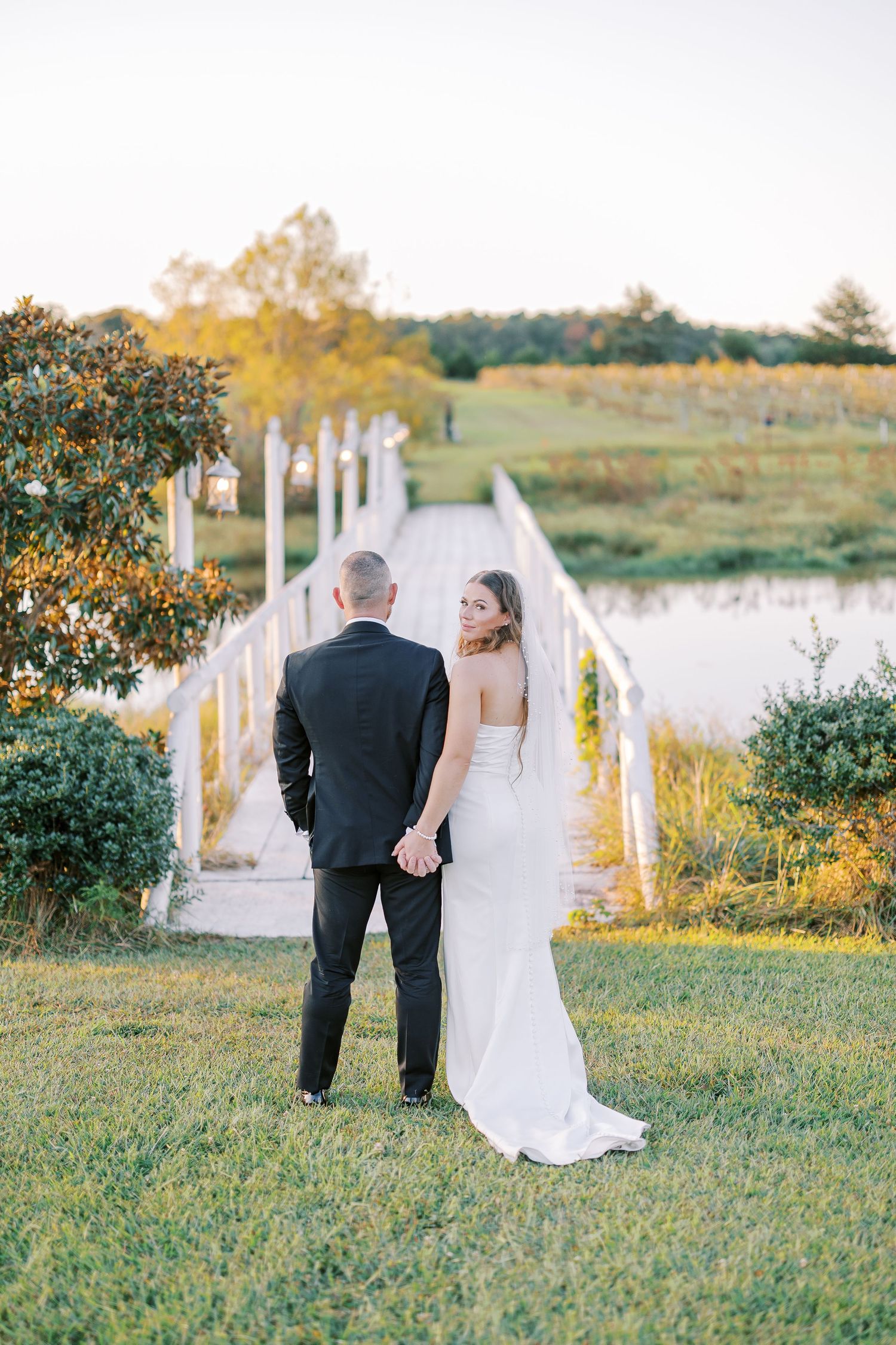 A wedding couple walks along a scenic outdoor path with white posts and golden sunlight in the background.