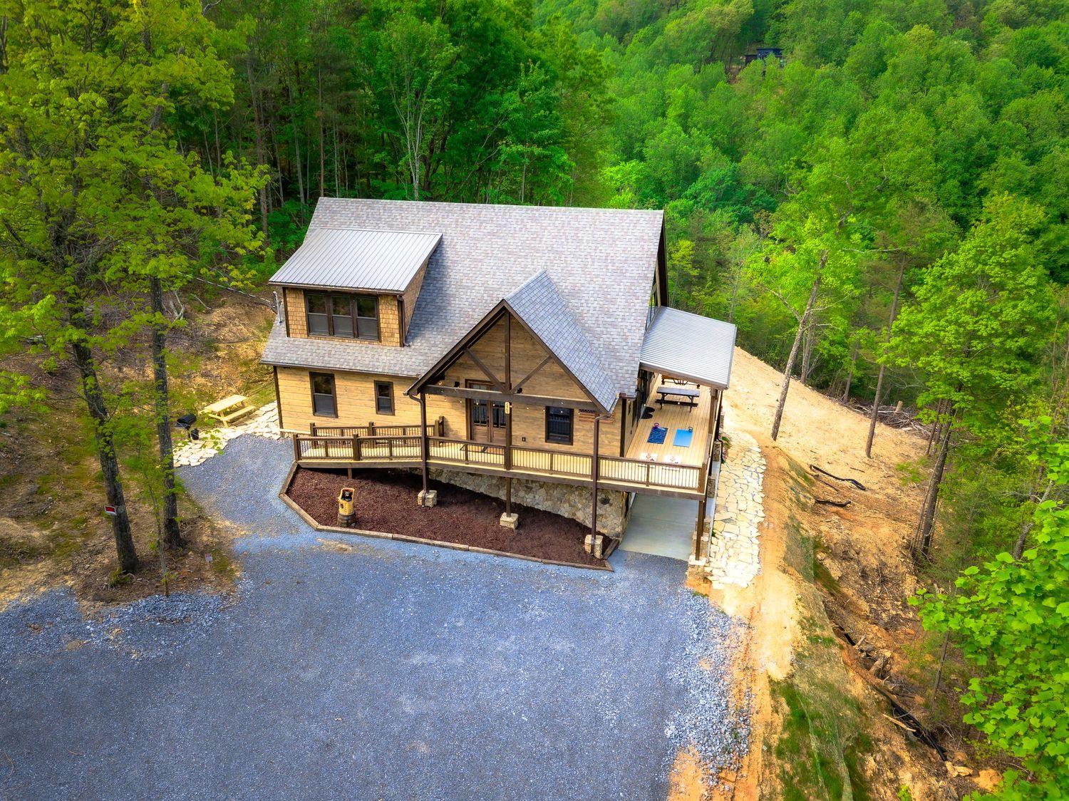Aerial view of a modern mountain cabin nestled among lush green forest with gravel driveway and parking area.
