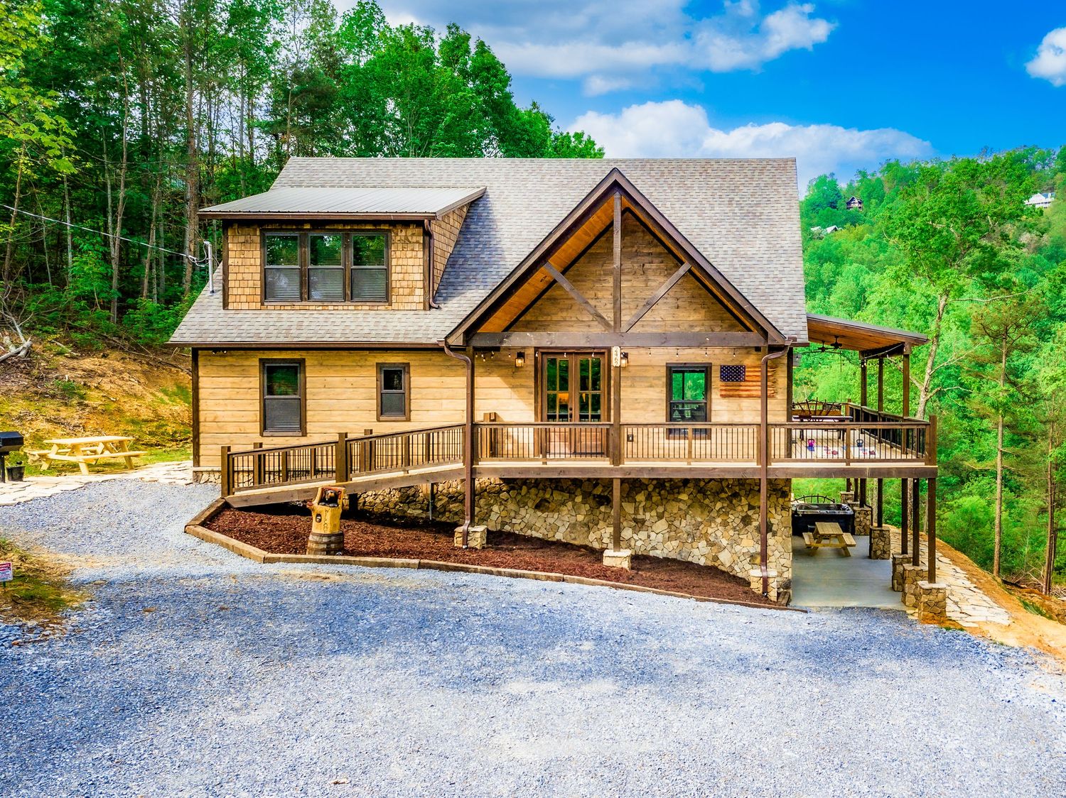 Modern rustic log cabin nestled in lush forest with gravel driveway and expansive deck surrounded by green trees.