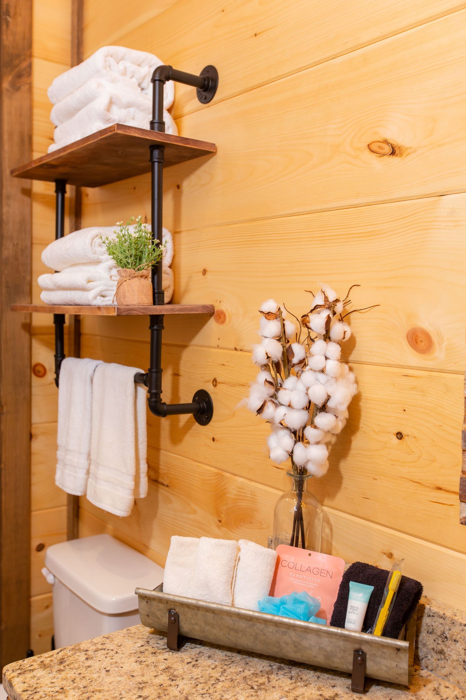Rustic bathroom interior with wooden walls, white towels on industrial pipe shelving, and cotton plant decorations.