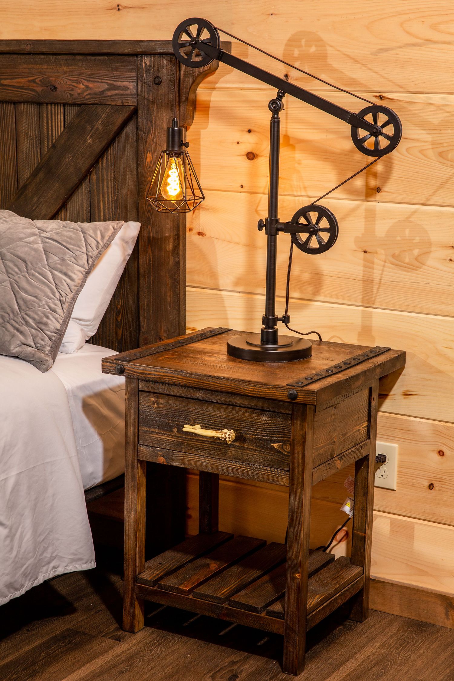 Industrial-style bedside table with vintage pulley lamp and wooden finish against pine walls.