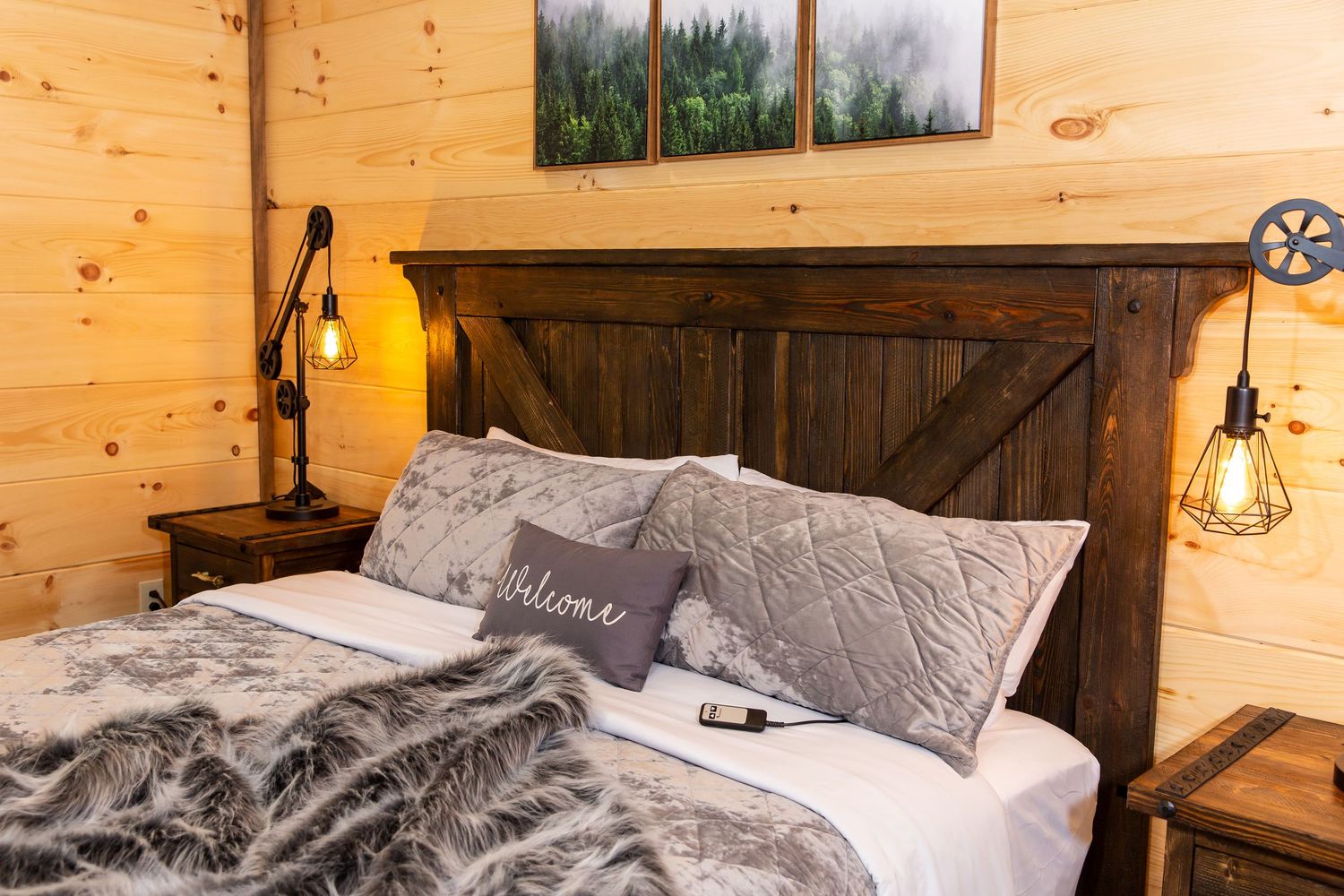 Cozy cabin bedroom with wooden walls and grey bedding overlooking forest views through large windows.
