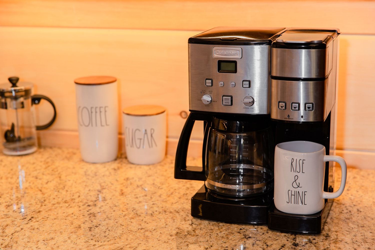 Coffee maker and matching white ceramic canisters arranged on a granite countertop in warm lighting.