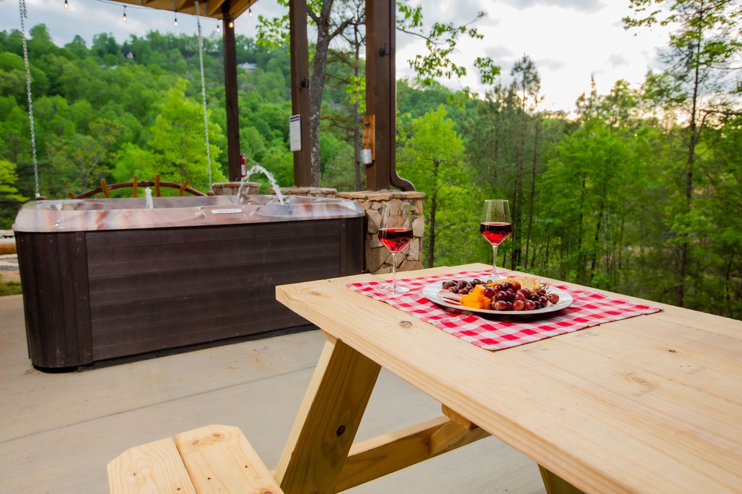 Rustic wooden picnic table with red checkered tablecloth and wine glasses on a covered outdoor deck overlooking forest views.