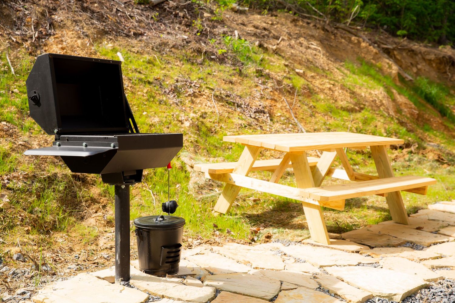 Wooden picnic tables with black grills arranged along a rocky hillside in a sunny outdoor setting.