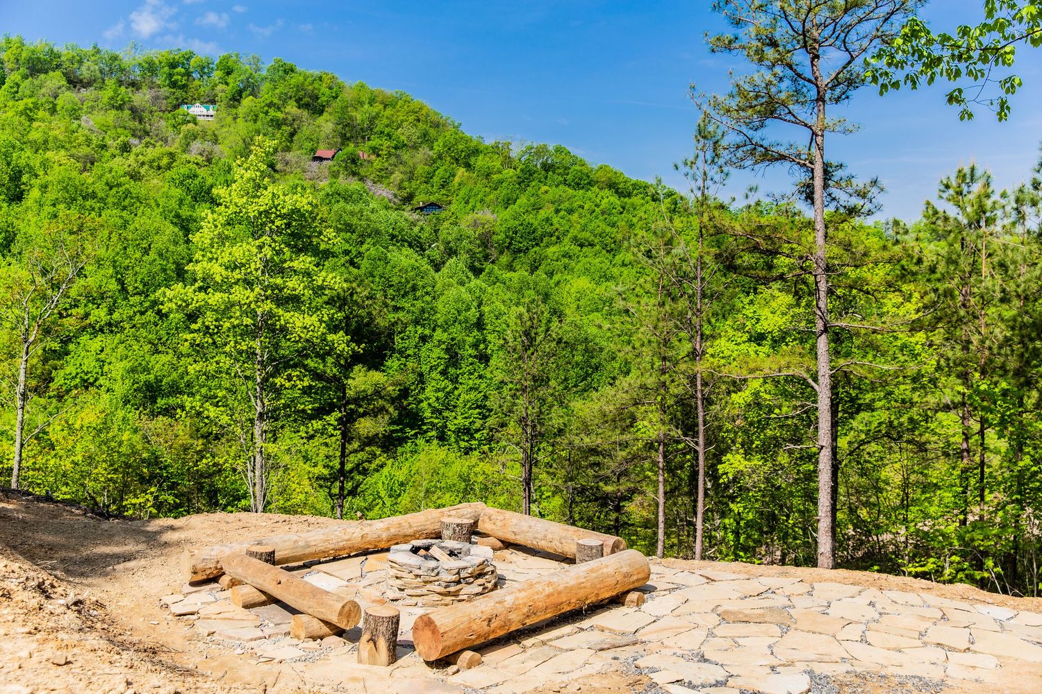 Scenic mountain landscape with cabin visible among green trees under bright blue sky.