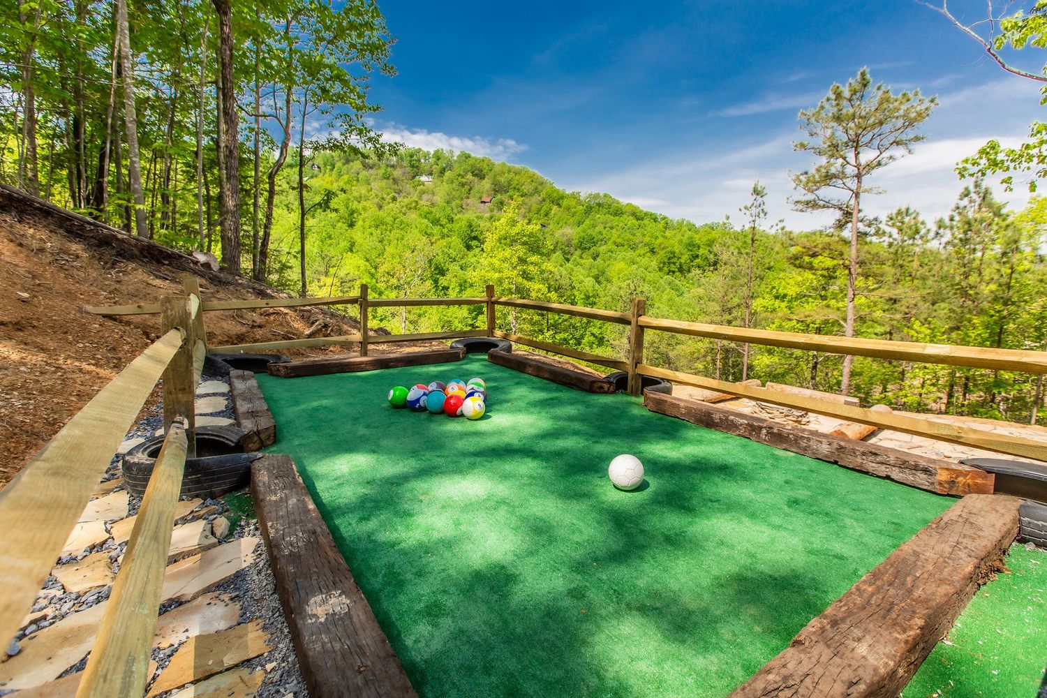Mini golf course with green turf and wooden borders set against a backdrop of mountain forest landscape.