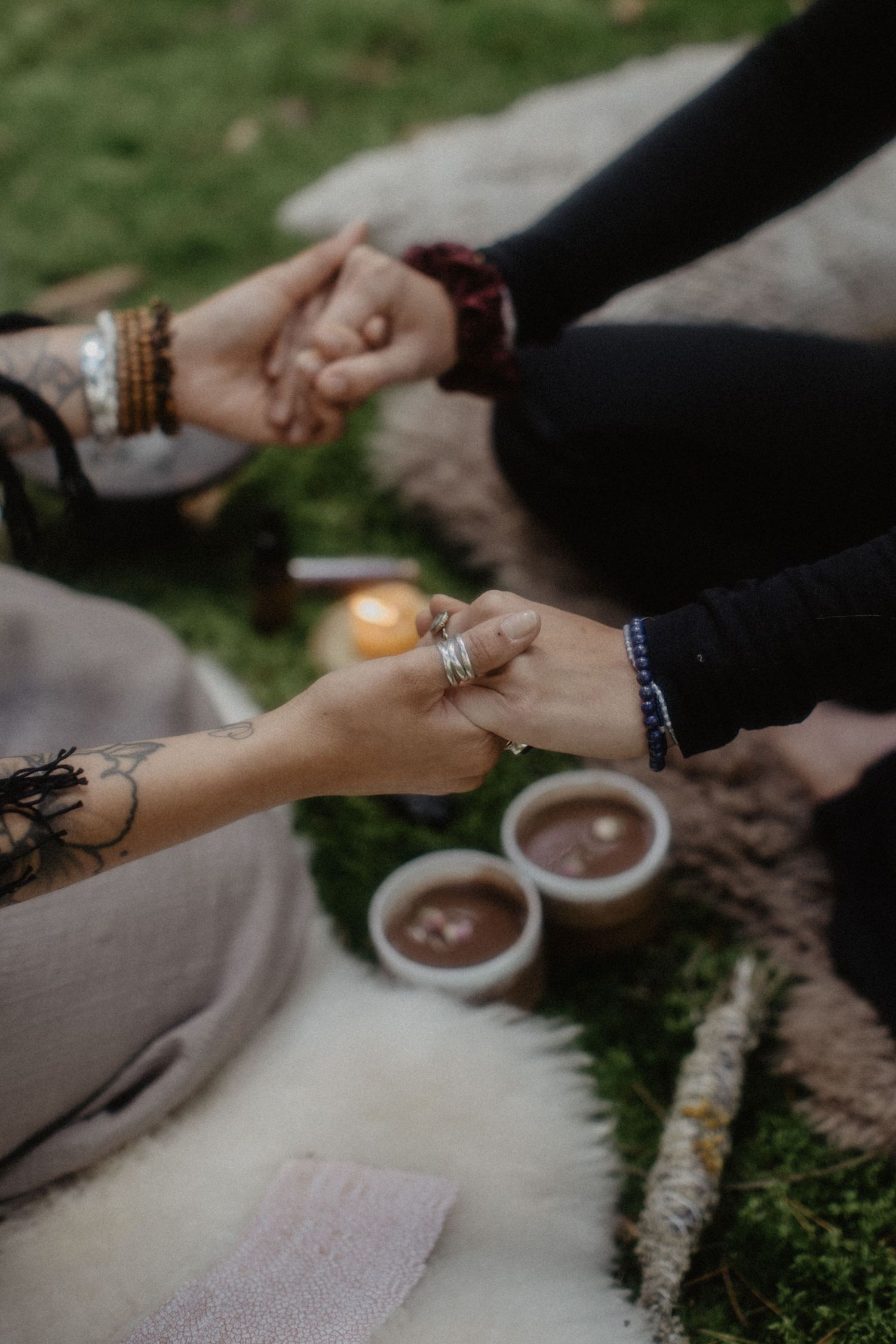 Hands hold cups of hot chocolate with marshmallows while sitting outdoors on a cozy blanket.